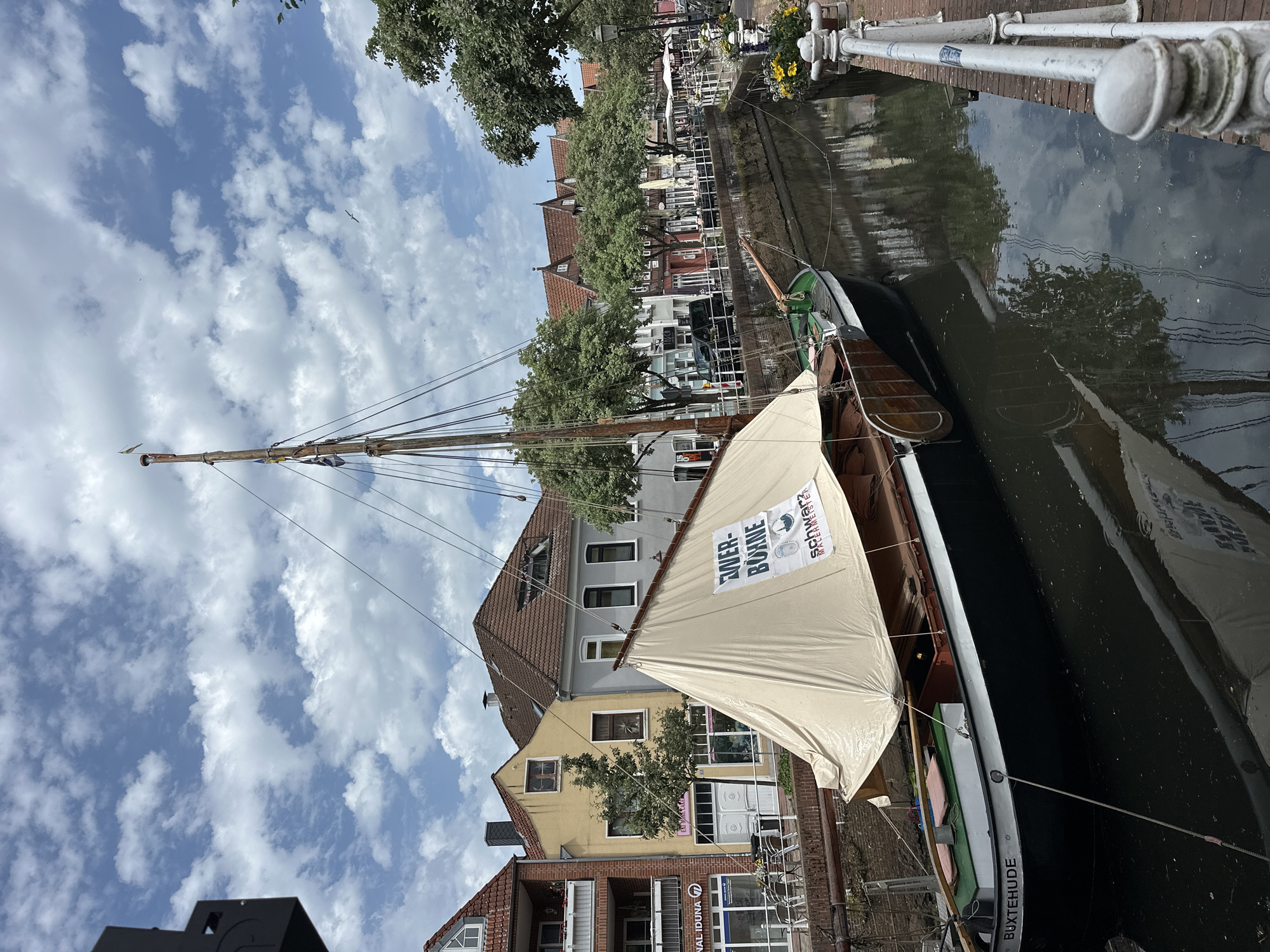 The image captures a scene in Buxtehude, Germany, featuring a docked historical sailing boat in a canal. The boat has a tall wooden mast and a light-colored sail with the words "Ewer-Bonne" and "Schwarzer Meerermeister" printed on it. The boat's hull is black and brown. The word "BUXTEHUDE" is inscribed on the side of the boat near the stern.
Buildings line the canal, including brick and painted houses with pitched roofs. Trees are interspersed along the canal bank, providing shade. A white fence separates the walkway from the canal. The sky is blue with scattered white clouds. The water reflects the buildings and sky.