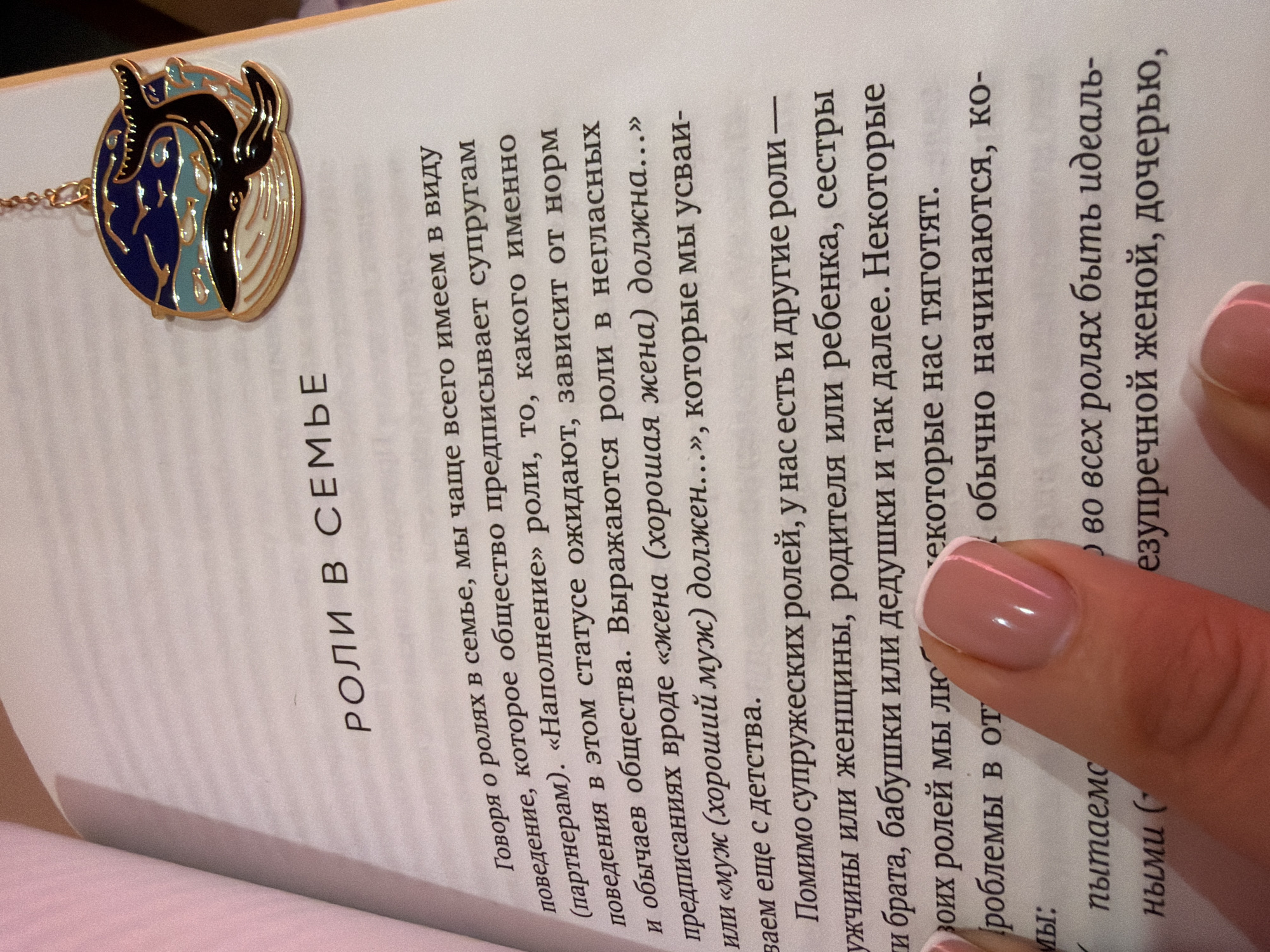 Here is a detailed description of the image:
The image is a close-up shot of a book page with Russian text on it, with a decorative necklace or bookmark laid on top of the page. The top of a hand with a freshly manicured nail is also visible, holding the page in place.
**People:**
- A hand and manicured nail
- Readers of the book
**Subjects:**
- Book: The book is open to a page titled "Roles in the Family" in Russian. The text discusses societal expectations for spouses and various family roles.
- Jewelry/Bookmark: The accessory looks like a round pendant or charm with a design featuring black and blue.
- Russian text.
**Objects:**
- Book
- Jewelry/Bookmark
- Hand/fingernails
**Actions:**
- Holding
- Reading
**Interactions:**
- A person is reading the book and using the decorative jewelry as a possible bookmark.
**Environment:**
- The scene appears indoors, likely in a home or quiet reading space, with soft, warm lighting. The background is blurred, indicating a shallow depth of field.
**Speculative Location**
The location cannot be verified.