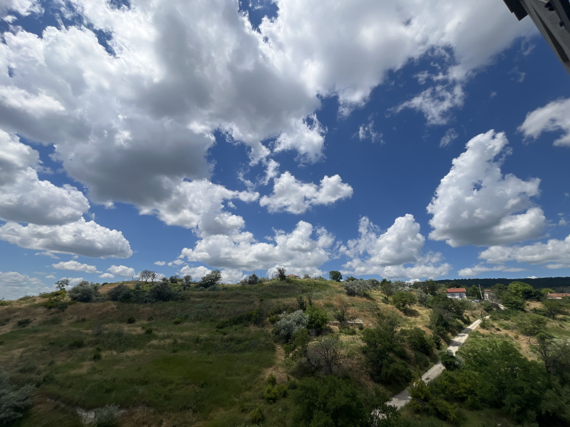 The image captures a scenic view of a hilly, vegetated landscape under a bright blue sky dotted with fluffy white clouds. The terrain is covered in dry, green grass and scattered trees and bushes, suggesting a Mediterranean or similar climate. A narrow, winding dirt path is visible, leading up the slope towards a cluster of buildings. The buildings appear to be houses with red-tiled roofs. The overall impression is one of a rural or semi-rural area on the outskirts of a city. Based on the characteristic topography and vegetation, this scene is likely located in the Varna region of Bulgaria.