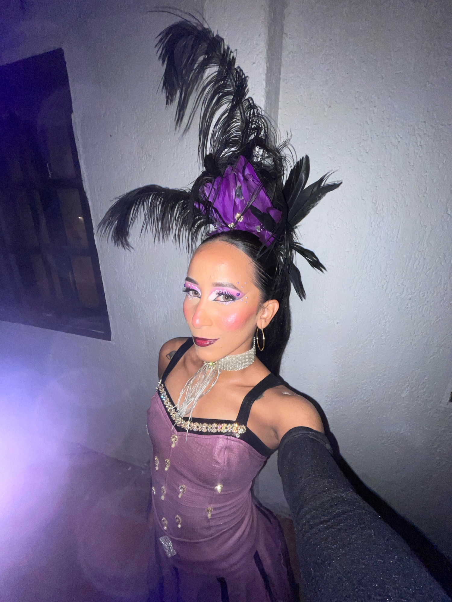 The image shows a person dressed in a theatrical costume. They are wearing a sparkly purple corset-style dress with beaded embellishments and black straps. A large feathered headdress, featuring purple and black feathers, is worn on their head. The person has dramatic makeup, including bold pink eyeshadow, glitter accents on their cheeks, and dark lipstick. They are also wearing a silver sequined choker necklace and a long black sparkly glove on their right arm. The background is a textured wall and a window with a dark frame. Purple light and mist are present on the left side of the image. The image appears to be a selfie taken at night, possibly in a dimly lit indoor location. No specific location in Porlamar, Venezuela is identifiable from the image.