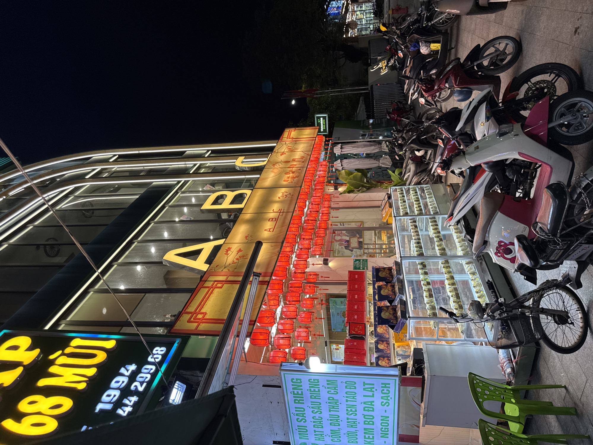 The image captures a nighttime scene on a street in Nha Trang, Vietnam, likely a commercial area. The focal point is a brightly lit building with a modern facade, adorned with large yellow letters spelling "A B C". The lower level of the building appears to be a shop, possibly a bakery or food establishment, given the display cases filled with items and the decorative red lanterns strung across the storefront.
A prominent green sign on the left displays text in Vietnamese, including "MÚI SẦU RIÊNG" and "HAT DAC SAU RIÊNG," indicating durian-related products. Below this, "KEM BƠ DA LAT" suggests avocado ice cream from Da Lat. Other phrases like "NGON - SACH" translate to "delicious - clean."
Several motorcycles are parked in front of the shop, indicating a common mode of transportation in the area. The street is paved, and the overall atmosphere is bustling, illuminated by the shop's lights and streetlights. The building's architecture suggests a contemporary design, with glass windows and metallic accents. The presence of outdoor seating with green chairs suggests a place where customers might dine or relax. The image is taken at eye level, offering a clear view of the shopfront and the activity around it.