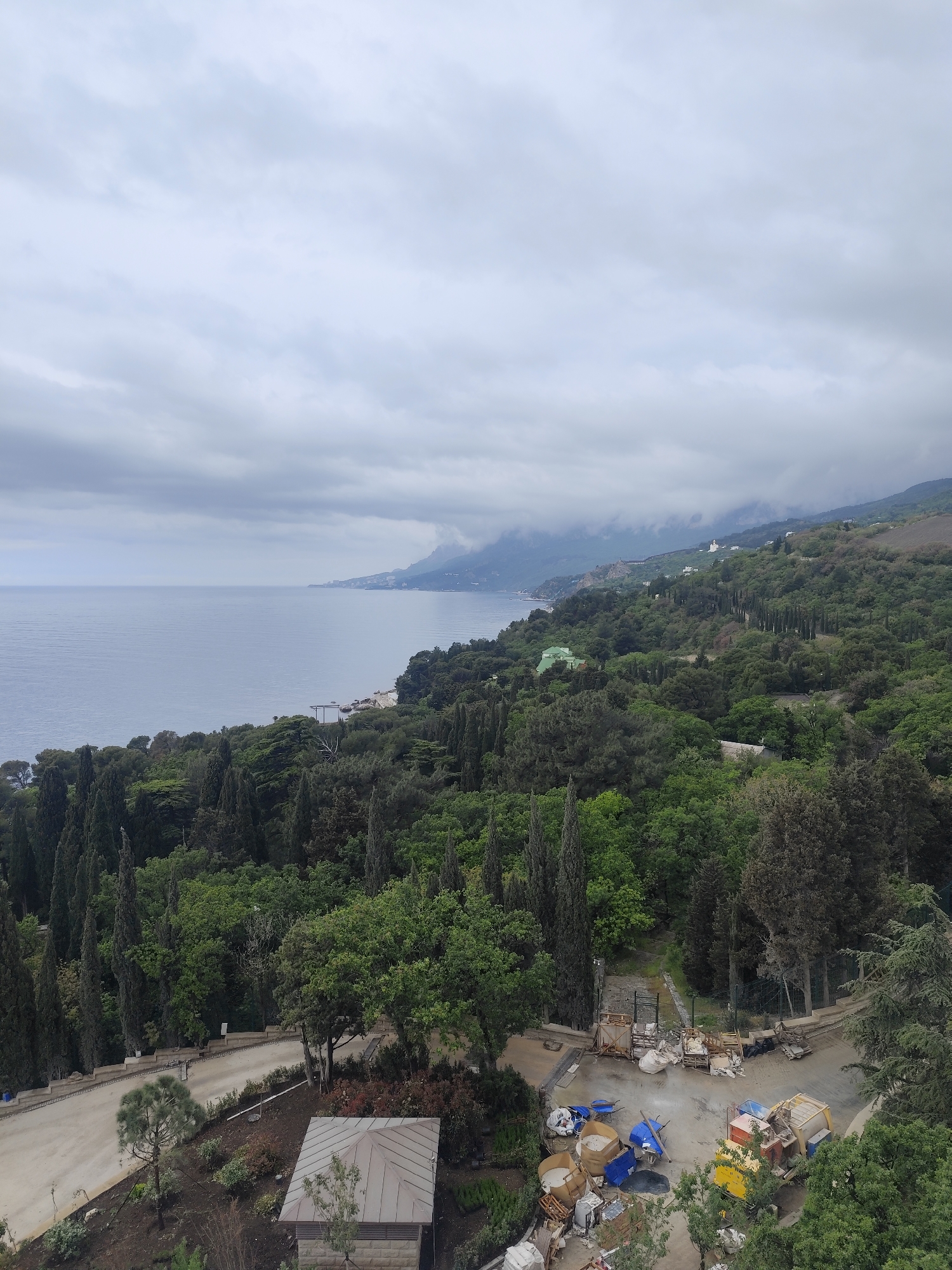 The image presents a view of a coastline under a cloudy sky. The water body, likely a sea or ocean, stretches to the left of the frame, meeting the horizon under the overcast sky. The shoreline is rugged, with a mix of greenery and buildings visible along its edge.
On the right, the landscape is dominated by a dense forest. Various shades of green indicate a diverse range of tree species. The terrain rises into hills or mountains, with some higher elevations obscured by clouds.
In the foreground, there is a road or pathway winding through the vegetation. A small building with a visible roof is present. A construction or dumping area is seen with various materials, equipment, and debris scattered around. There are containers, machinery, and piles of what appears to be construction waste.
The scene suggests a coastal environment undergoing some form of construction or maintenance, possibly in a hilly or mountainous region.