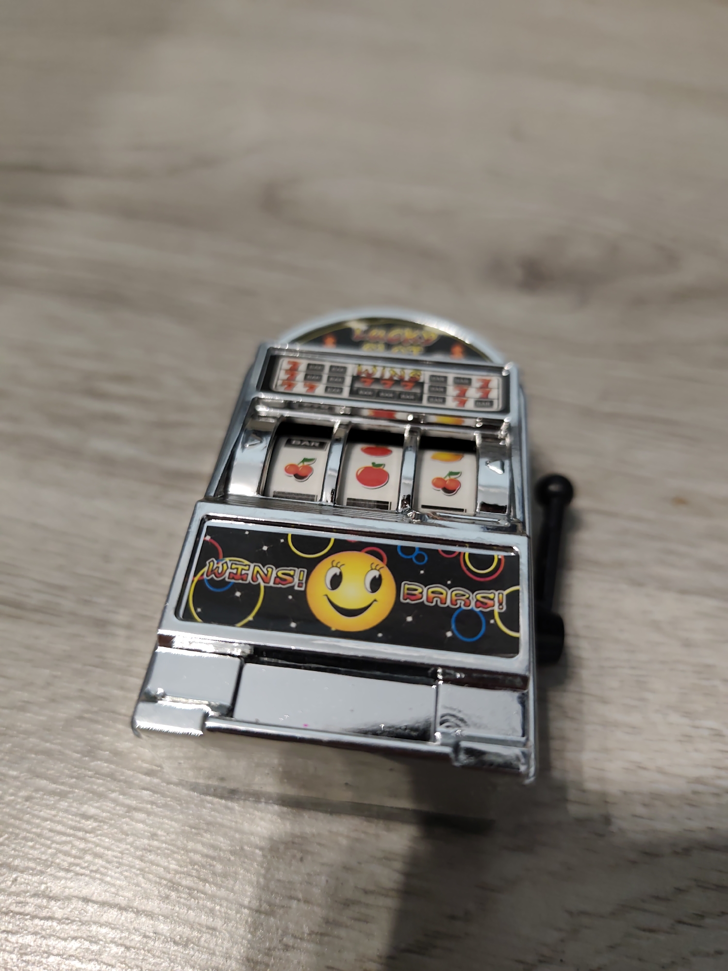 The image displays a miniature, chrome-finished slot machine resting on a light brown wooden surface. The location, Pazardzhik, Bulgaria, cannot be visually confirmed from the image itself.
The slot machine is the central subject, featuring three internal reels. From left to right, these reels display a cherry, an apple, and a cherry symbol. Above the reels, obscured text includes "BAR" and "777". Below the reels, a black panel features the yellow text "WINS!" on the left and "BARS!" on the right, both with red/orange outlines. Between these words, a yellow smiley face with rosy cheeks and eyelashes is prominently displayed. The black panel's background is patterned with various colored rings and circles (blue, yellow, red). A black lever, characteristic of a slot machine, extends from the right side.
The wooden surface beneath the slot machine exhibits a visible grain pattern, suggesting either wood or laminate. The background is out of focus, showing a blurred continuation of the same wooden surface. A distinct shadow of the slot machine is cast towards the lower right of the frame, indicating a light source from the upper left. The chrome surfaces of the machine show reflections. No people are present in the image.