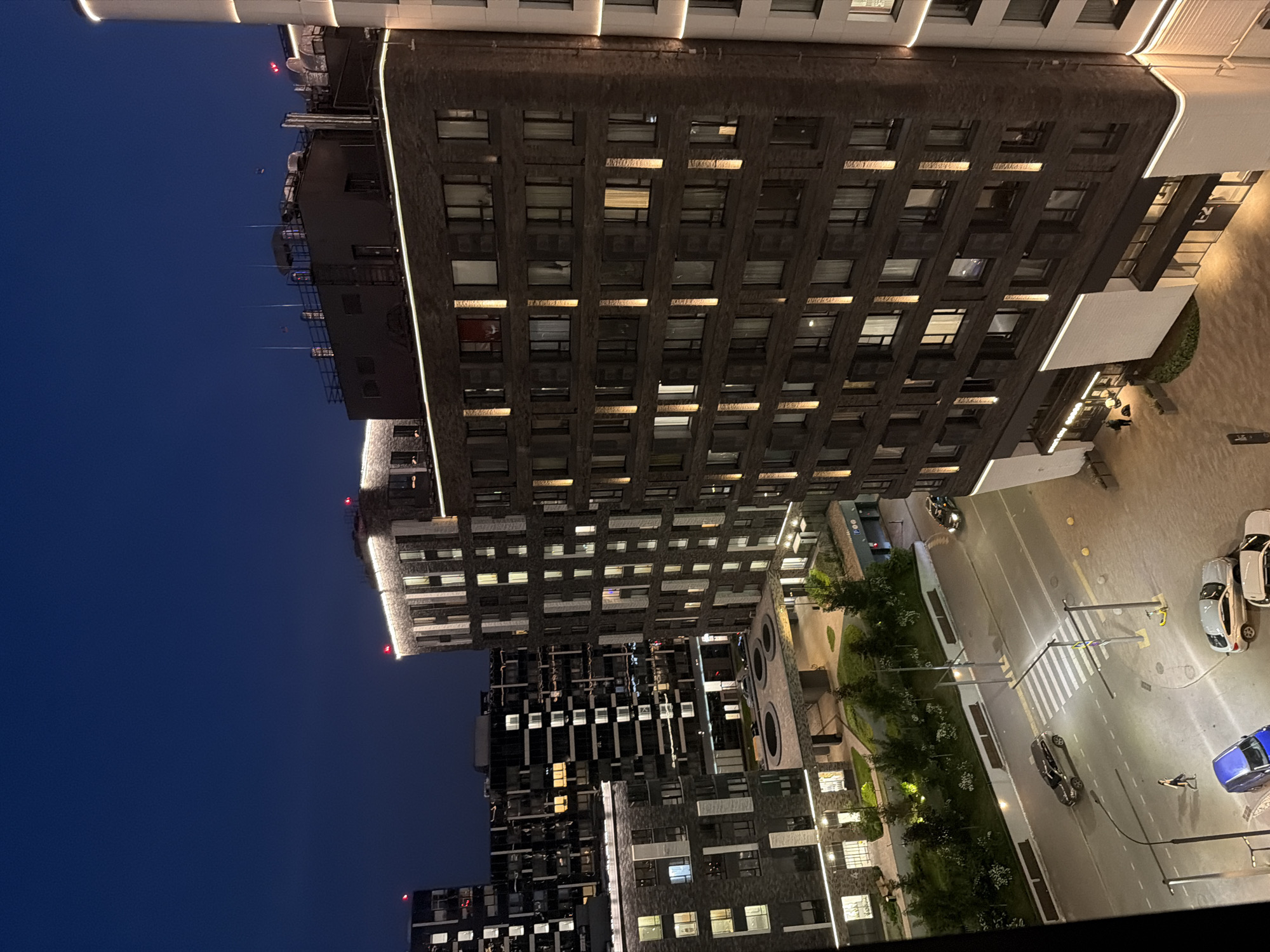 An elevated, nighttime view captures a modern urban residential complex, likely in Moscow, Russia, given the architectural style and building density. The scene is dominated by several multi-story apartment buildings constructed with dark brick and featuring prominent, illuminated vertical strips and horizontal bands of light. Many windows are lit, indicating occupancy.
In the foreground, a street runs through the complex, with a pedestrian crosswalk clearly visible. A few cars are parked and in motion on the street. One car, a dark SUV, is driving across the crosswalk. A woman is walking on the sidewalk to the left of the crosswalk. A bright blue van is parked on the left side of the street, and a silver sedan and a white sedan are parked on the right. A yellow scooter is parked near a street lamp on the right side of the road.
The buildings are interconnected by elevated walkways and courtyards, which are landscaped with trees and low bushes. Some buildings have ground-level commercial spaces with illuminated storefronts. The overall atmosphere is one of a well-lit, active residential area at dusk or early night. The sky is a deep, clear blue, indicative of twilight or a cloudless night.