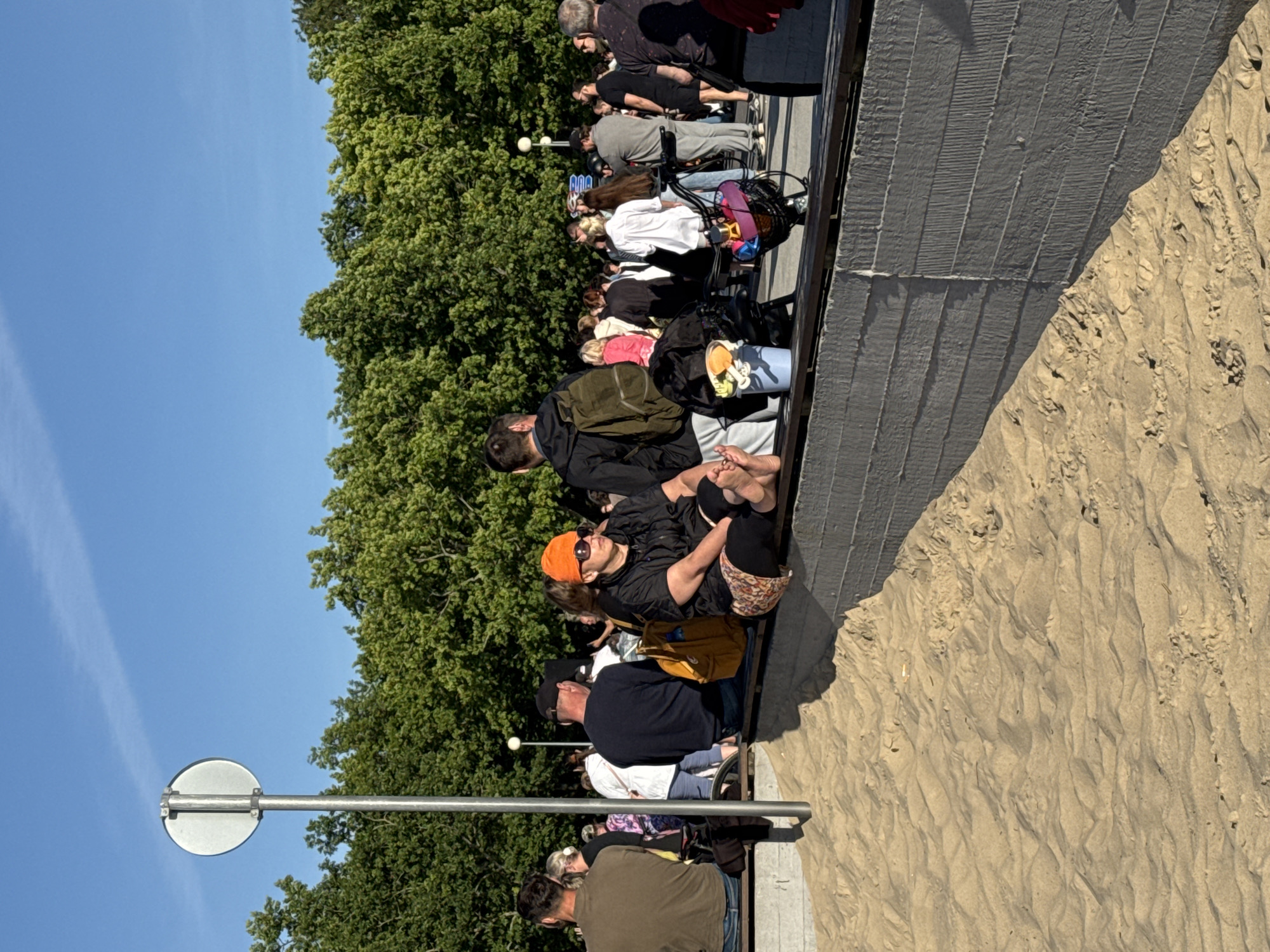 The image depicts a sunny outdoor scene at the Tallinn Song Festival Grounds in Tallinn, Estonia, a location known for large public gatherings. In the foreground, a sandy area is visible with textured patterns. A gray concrete wall with horizontal indentations runs along the edge of the sandy area, topped by a wooden railing.
Seated on the wooden railing in the mid-ground is a woman wearing an orange beanie, sunglasses, and a black jacket over a patterned skirt. Her bare feet are visible as she sits with her legs extended. To her left, a yellow backpack rests on the railing. Behind her, a crowd of people is gathered, some standing and some sitting on benches or the railing. The crowd is diverse in age and attire, with individuals holding bags and some with backpacks. Trees with lush green foliage form a backdrop to the scene. A metal pole with a circular sign is visible on the left side of the frame. The sky is clear and blue.