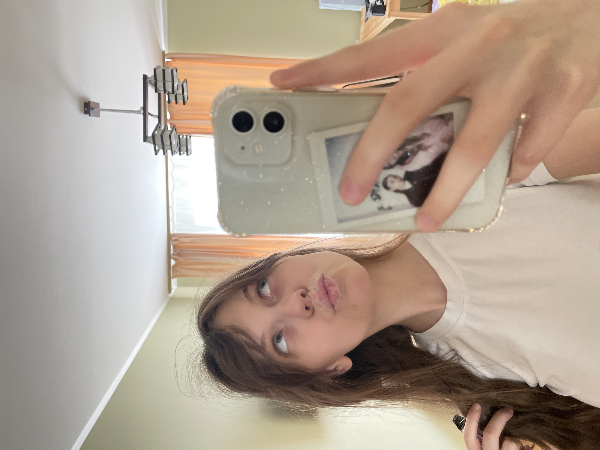 The media file is a selfie taken by a young person in what appears to be a bedroom. The person is making a duck face and looking up and to the left, with their hair cascading down. They are wearing a white t-shirt. The phone used for the selfie is visible, featuring a light-colored, glittery case with two small framed pictures. In the background, a wall with a light green paint color is visible, along with a window with orange blinds. A modern-looking wall-mounted light fixture with multiple lamps is also visible on the wall. The overall lighting suggests it is daytime. The image is oriented vertically, implying it was taken on a mobile device.