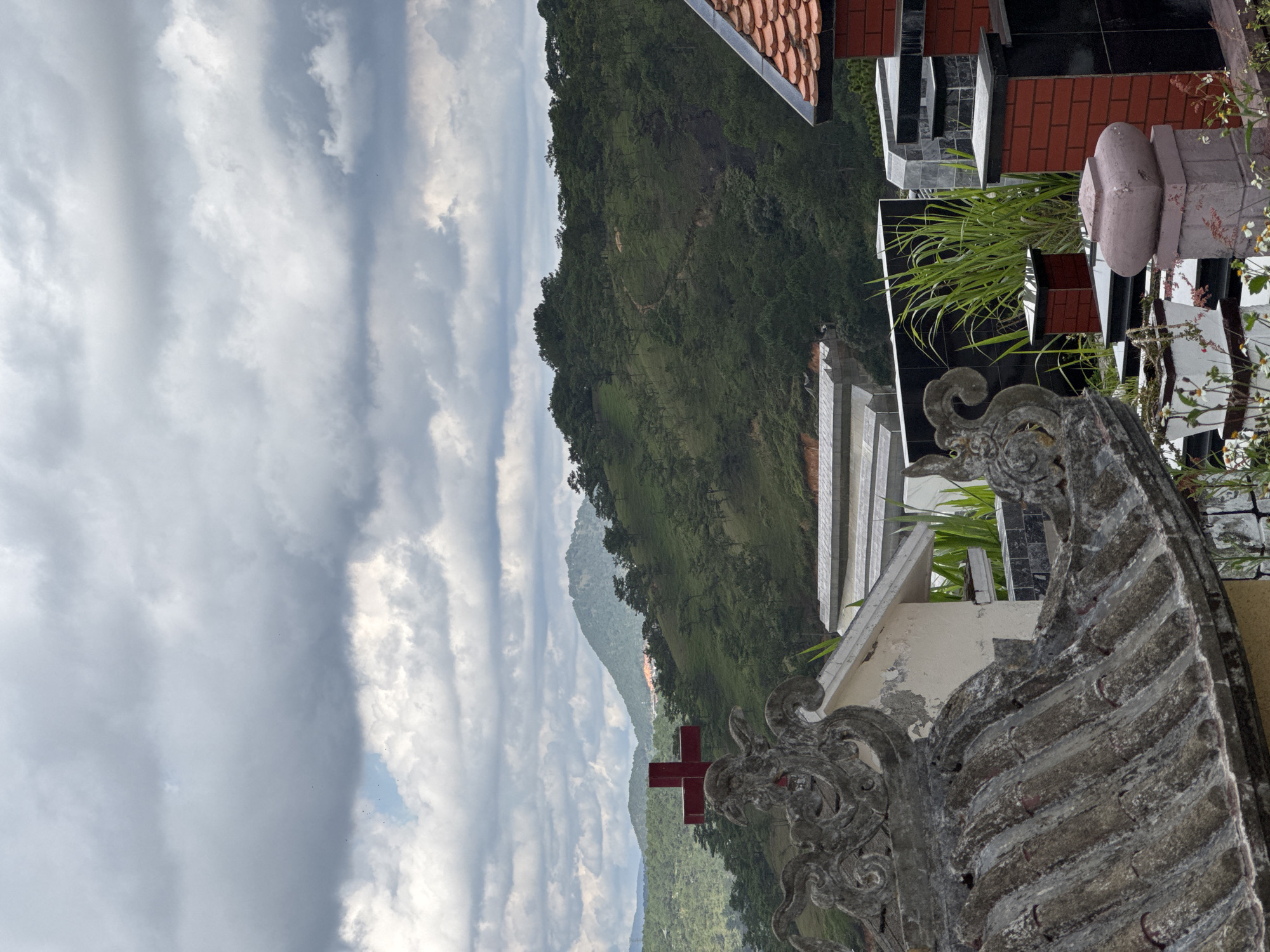 The image captures a view from a cemetery in Lạc Dương, Vietnam, looking out towards a mountain range under a cloudy sky. Foreground elements include intricately carved stone structures, typical of Vietnamese cemeteries, with a red cross visible in the distance. The graves are composed of brick, stone, and tile. Green foliage grows both near the grave sites and on the surrounding mountain. The cloudy sky provides a soft, diffused light to the scene.