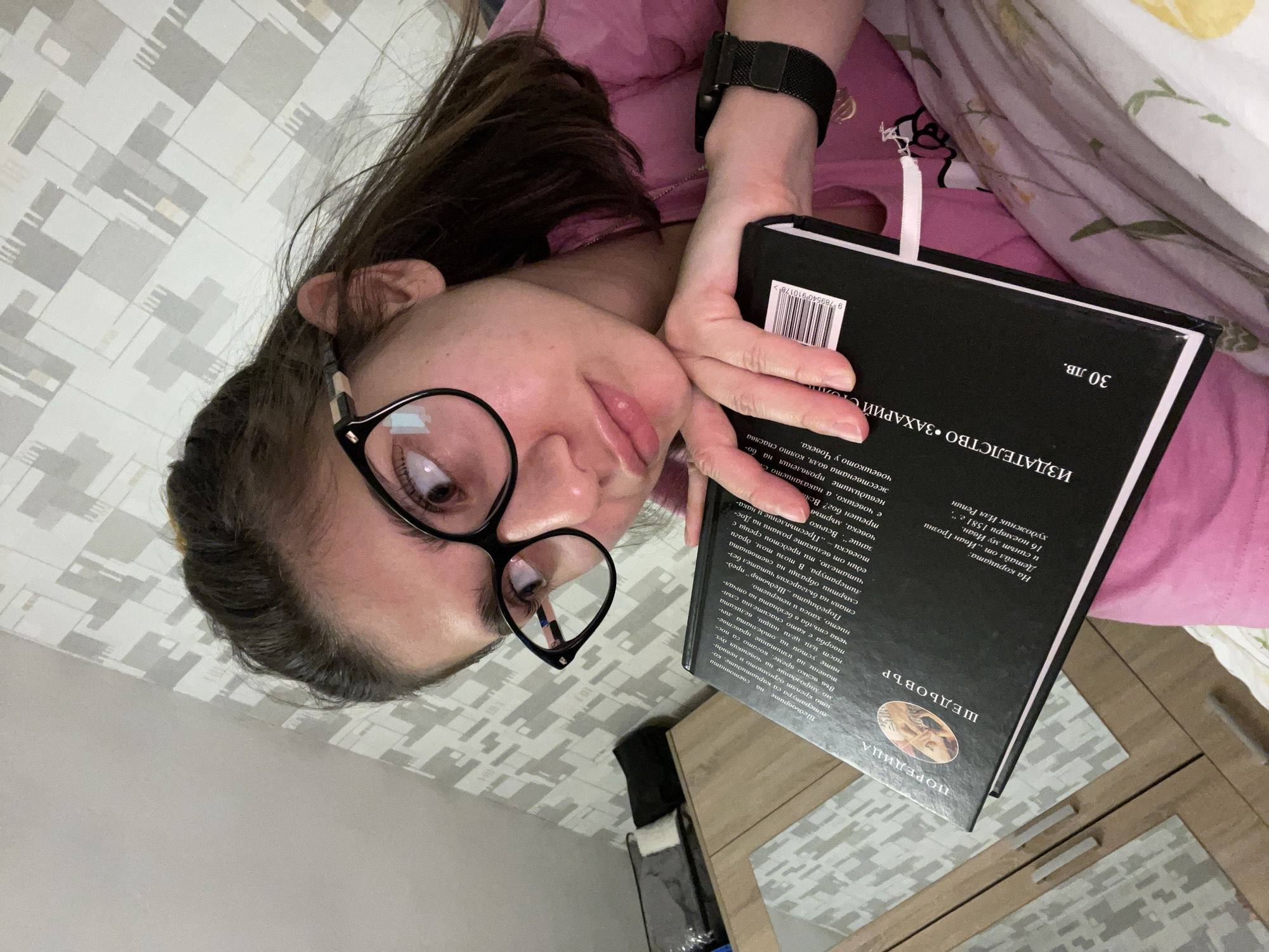 A woman with dark hair and glasses is lying on a patterned surface, possibly a bed or a couch. She is holding a book with a black cover, written in Bulgarian, and is looking at the camera with a slight smile. She is wearing a pink shirt and a black smartwatch. In the background, there is a wooden cabinet. Based on the text in the book, it is likely that this image was taken in Sofia, Bulgaria.