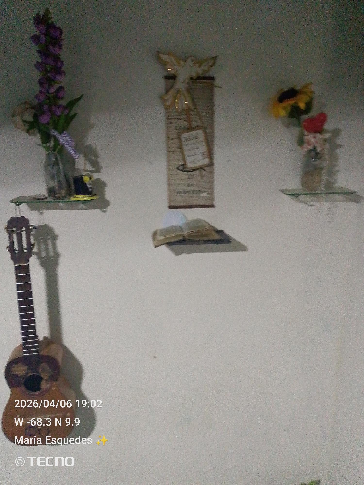 The image shows a wall with several objects displayed on it. On the left, there is a small ukulele hanging near a glass shelf with a vase of purple flowers. In the center, there is a decorative religious piece with a dove and a small open book on the wall below it. On the right, another glass shelf holds a vase with sunflowers and a red flower. The text overlay indicates the location as Tinaquillo, Venezuela, and the date as April 6, 2026. The text also includes the name María Esquedas and the camera brand, TECNO.