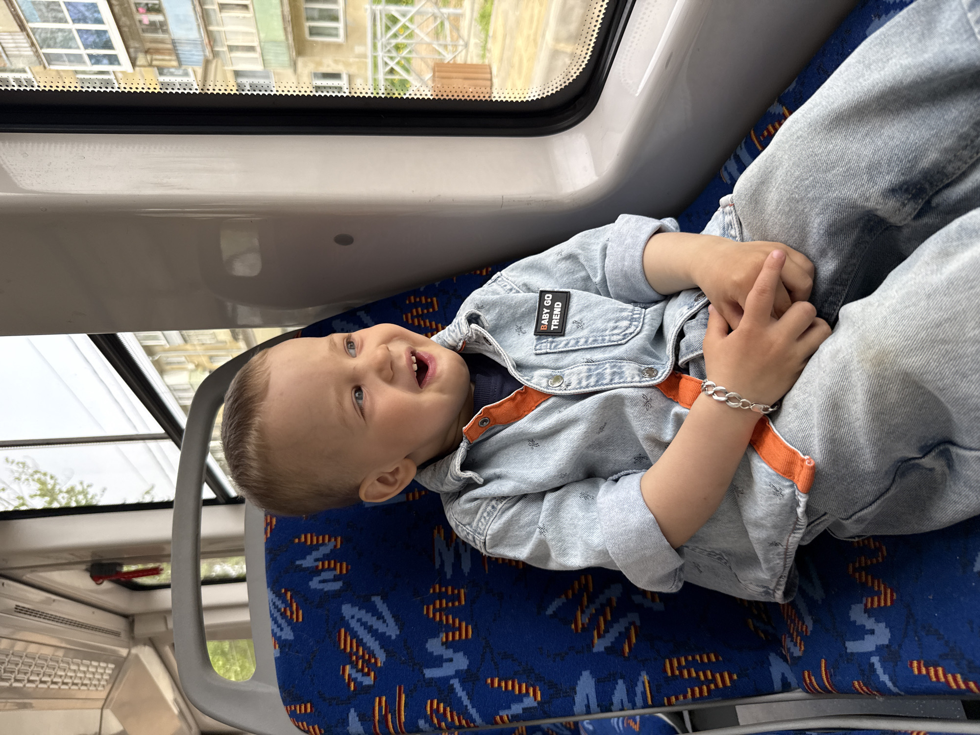 A small child lies on a blue patterned seat in what appears to be a train or bus. He is wearing a light blue denim jacket over an orange-collared shirt, and light blue pants. He has a silver bracelet on his wrist. The child is smiling and looking upwards towards the window. Outside the window, buildings are visible, consistent with an urban environment. Given the context and additional location information, the buildings are likely in the Rusanovo district of Russia. The interior of the vehicle is brightly lit.