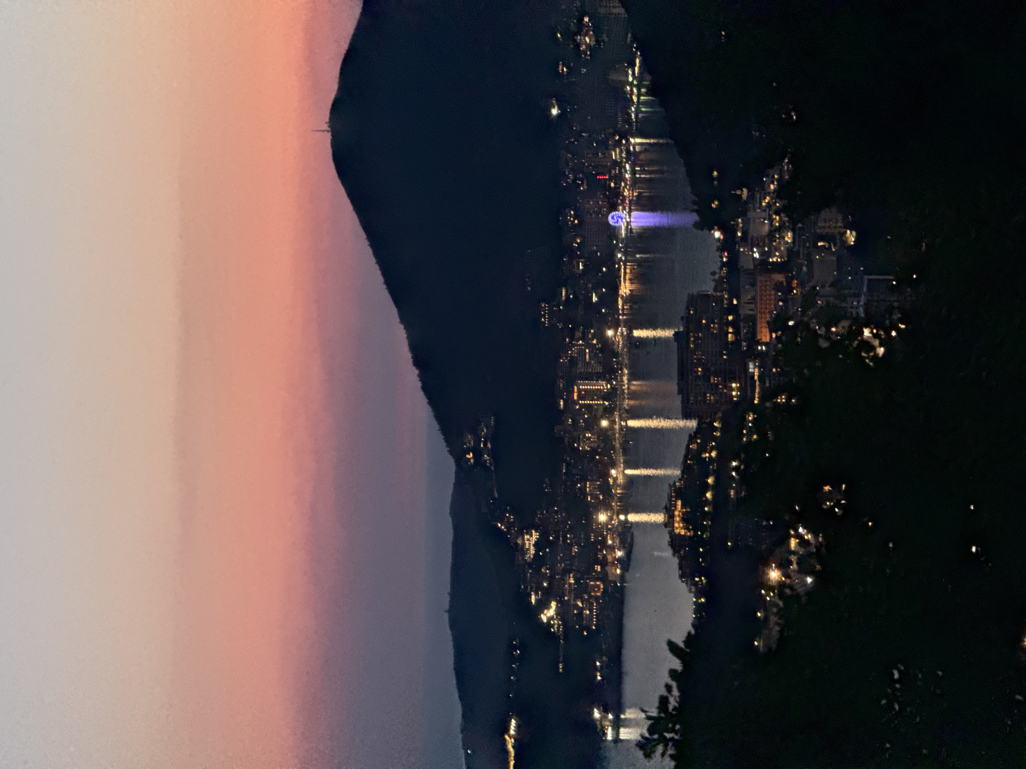 The image depicts a twilight view of the coastal city of Podbabac, Montenegro, illuminated by numerous lights. The sky displays a gradient of soft pink and purple hues, transitioning into a darker blue towards the horizon. In the foreground, dark, silhouetted foliage partially obscures the view.
The midground is dominated by the city spread along the coastline, nestled against rolling hills. Buildings of varying sizes are visible, many with illuminated windows and facades, creating a sparkling effect. A body of water, likely a bay or the sea, lies between the city and the observer, reflecting the lights from the shore. A Ferris wheel, glowing with bright blue light, stands out prominently on the waterfront. Several boats are moored or navigating the water, their lights also visible.
The background features a large, dark silhouette of a mountain, with a small structure, possibly a communication tower, visible at its peak. The overall impression is a serene and picturesque evening scene in a coastal urban environment.