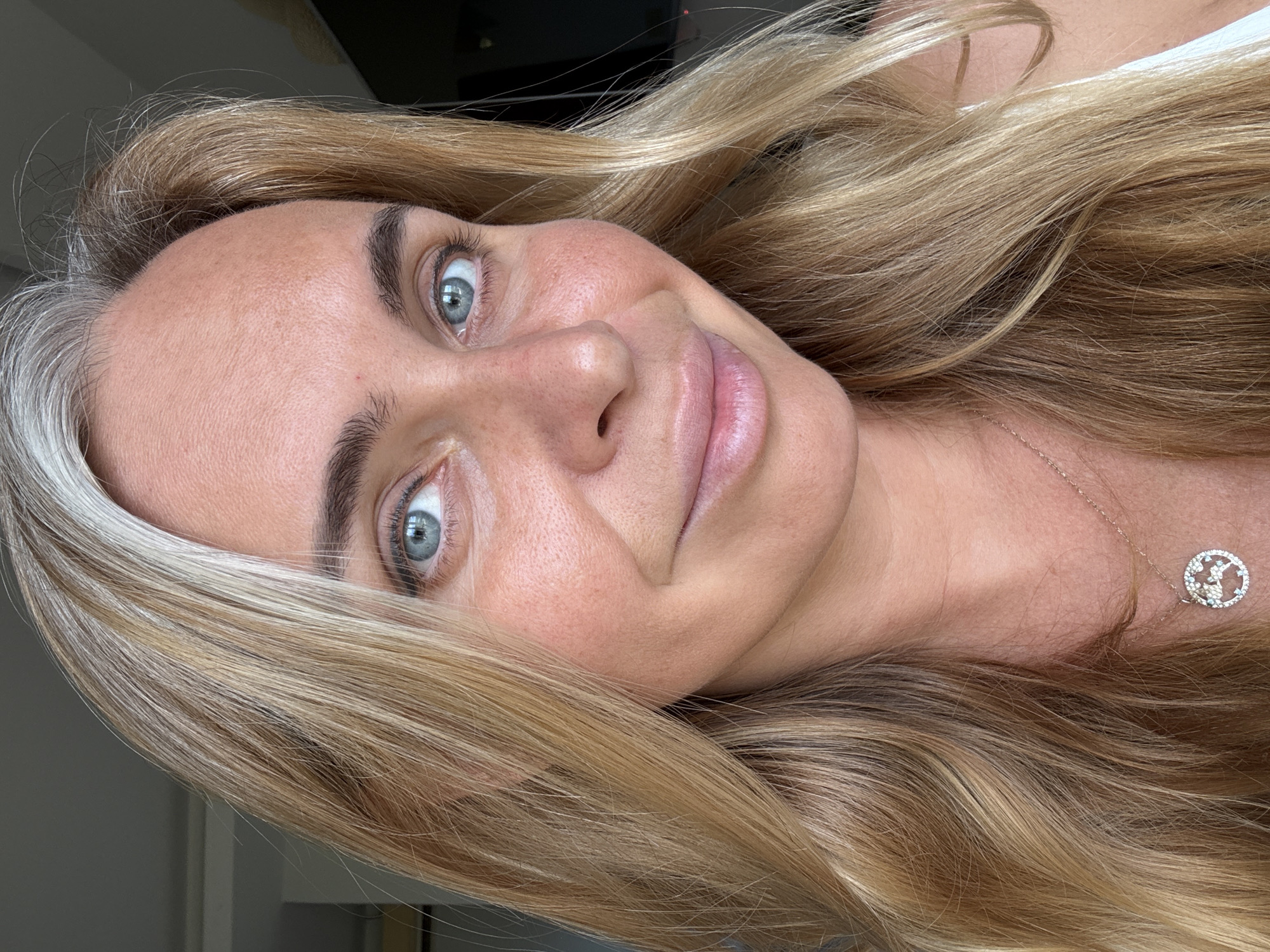 The image is a close-up portrait of a woman with fair skin, freckles, and blue eyes. Her blonde hair is styled in loose waves and falls around her face. She is wearing a delicate necklace with a circular pendant. The background is blurred, but appears to be indoors with natural light. There is no discernible information within the image to indicate the location in Baan Chaweng, Thailand.