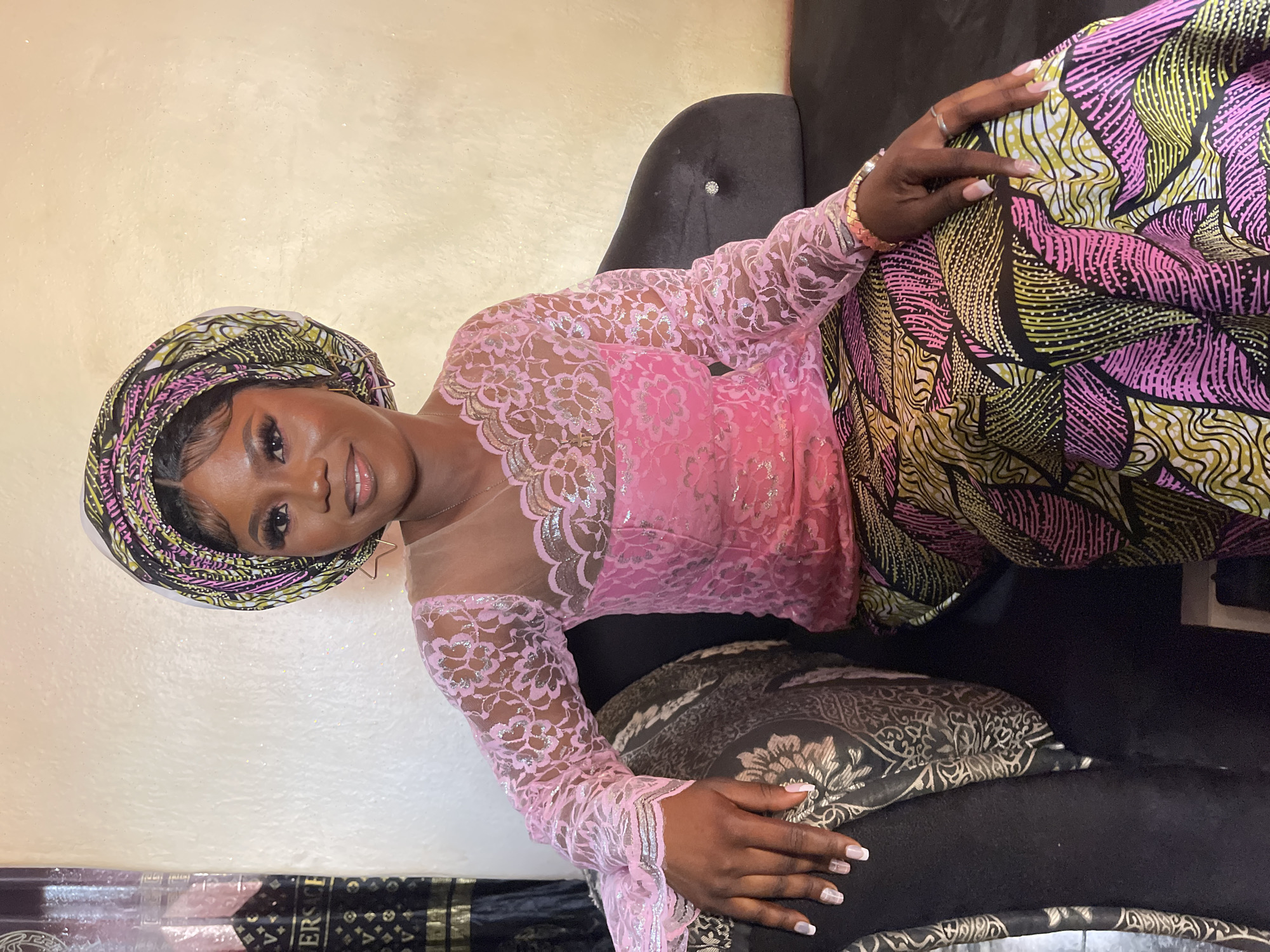 A young Black woman is pictured indoors, seated and smiling warmly at the camera in Akwanga, Nigeria. She is dressed in traditional Nigerian attire, featuring a vibrant, multi-colored headwrap (gele) with patterns in black, yellow-gold, pink, and white. Her top is a delicate pink lace blouse with long sleeves, adorned with metallic silver or light gold embellishments and a sheer, flesh-toned neckline. Complementing the gele, she wears a matching patterned wrapper or skirt in black, yellow-gold, and pink.
She is seated on a black upholstered armchair or sofa, with her hands gently resting on her lap and the patterned fabric. She wears several rings on her left hand and a bracelet on her left wrist, along with small, gold star-shaped earrings. Her fingernails are neatly manicured with white tips. The background is a light, textured wall, possibly with a subtle glitter finish, suggesting an interior setting with soft lighting. The overall impression is one of elegance and festive attire, suitable for a special occasion or portrait. Partially visible in the bottom left corner of the main image, a dark-colored item with the text "VERSACE" is discernible.