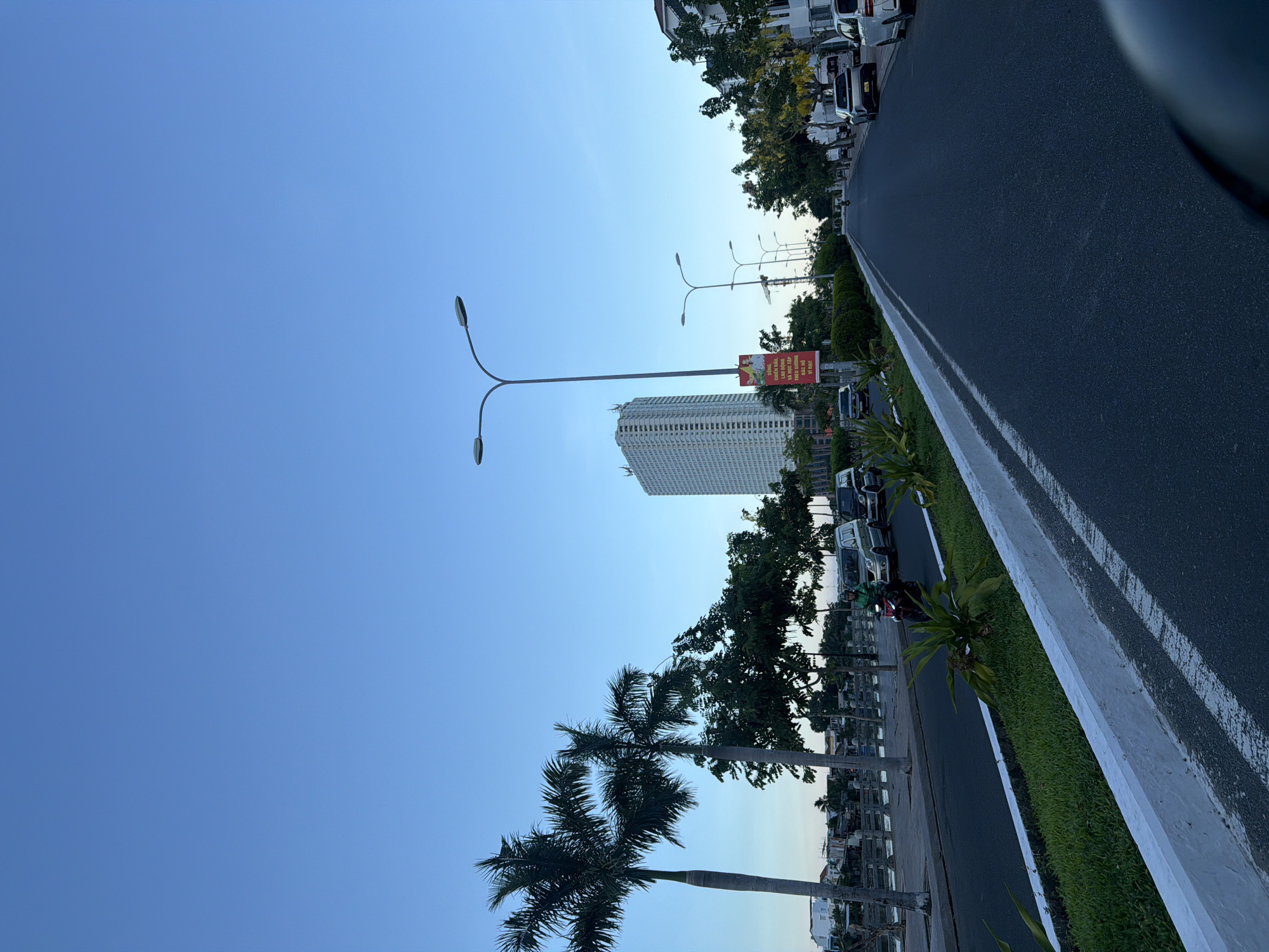 This image shows a street scene in Nha Trang, Vietnam, likely in the city or a coastal ward. The sky is a clear, light blue, suggesting early morning or late afternoon. On the left side of the road, palm trees and other lush greenery line a walkway next to a body of water, possibly a canal or part of the coastline. Several vehicles, including cars and a motorcycle with a rider, are present on the asphalt road. A tall, white, modern building dominates the skyline on the left, while residential buildings and more trees are visible on the right. A red banner with Vietnamese text is displayed on a pole near the road. The overall impression is of a well-maintained urban environment with abundant vegetation.