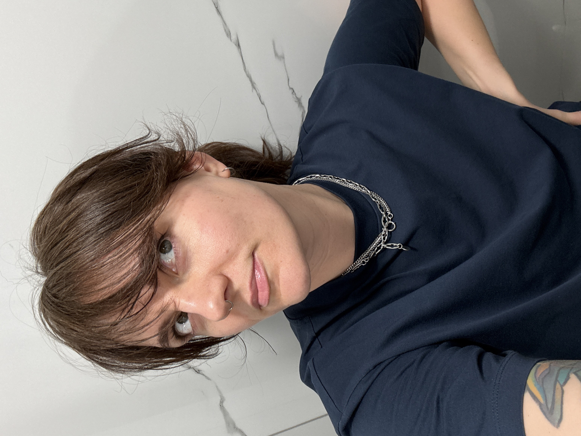 A medium shot depicts a person with fair skin, brown hair, and a nose ring. They are wearing a dark blue T-shirt and a multi-layered silver chain necklace. Part of a tattoo is visible on their left arm. The background is a white surface with dark grey marbling. The location is undetermined; there is no indication of Budva, Montenegro.