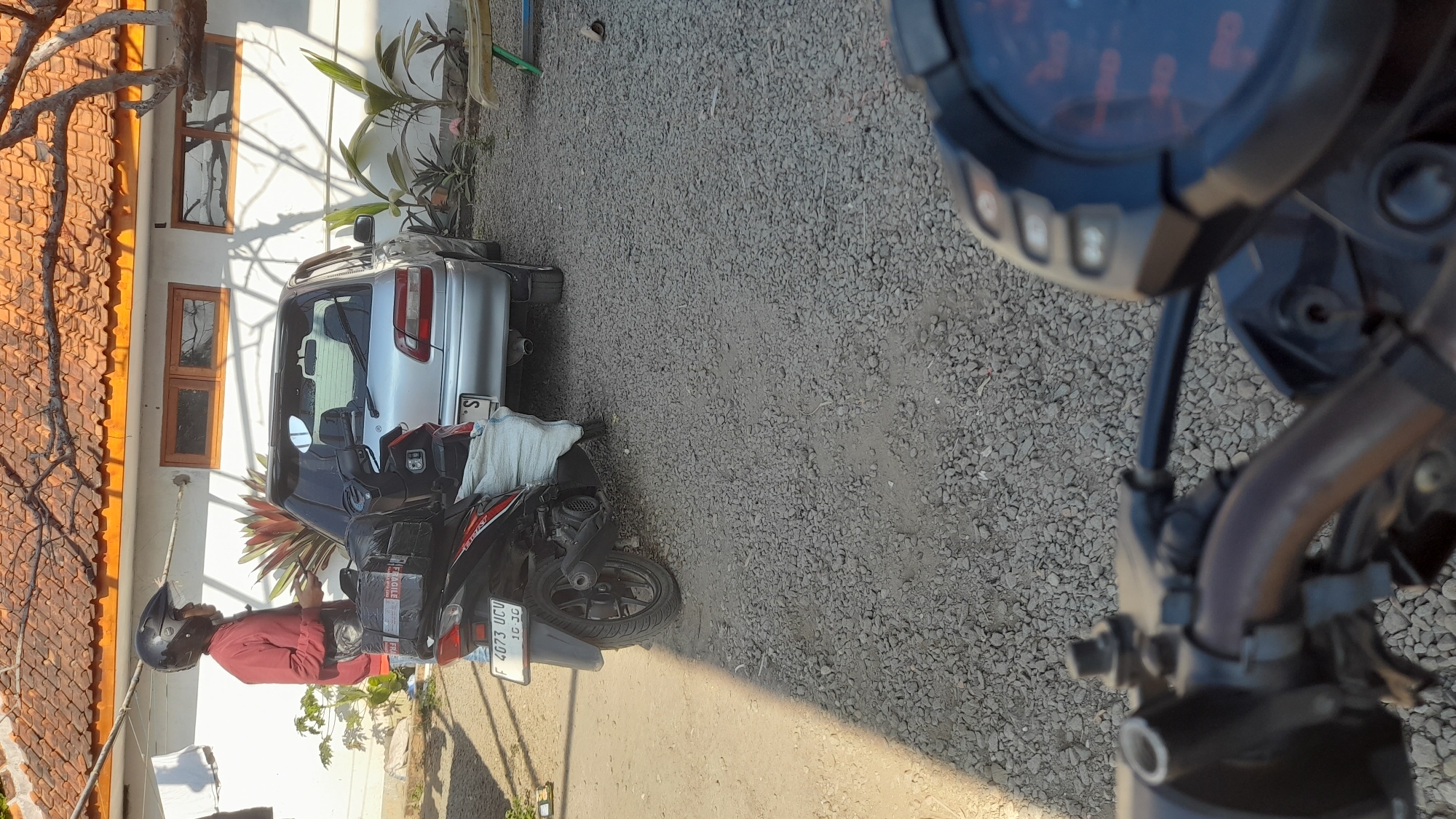 In the city of Selaawi, Indonesia, a man wearing a red jacket and a black helmet stands next to a silver sedan and a black motorcycle. The motorcycle has a package strapped to its rear, and a white bag is also attached. The man is holding a smartphone. The scene appears to be a residential driveway, with a house in the background that has an orange-tiled roof and white walls with wooden-framed windows. The ground is covered in gravel. The lighting suggests it is daytime, with shadows cast by trees and the house.