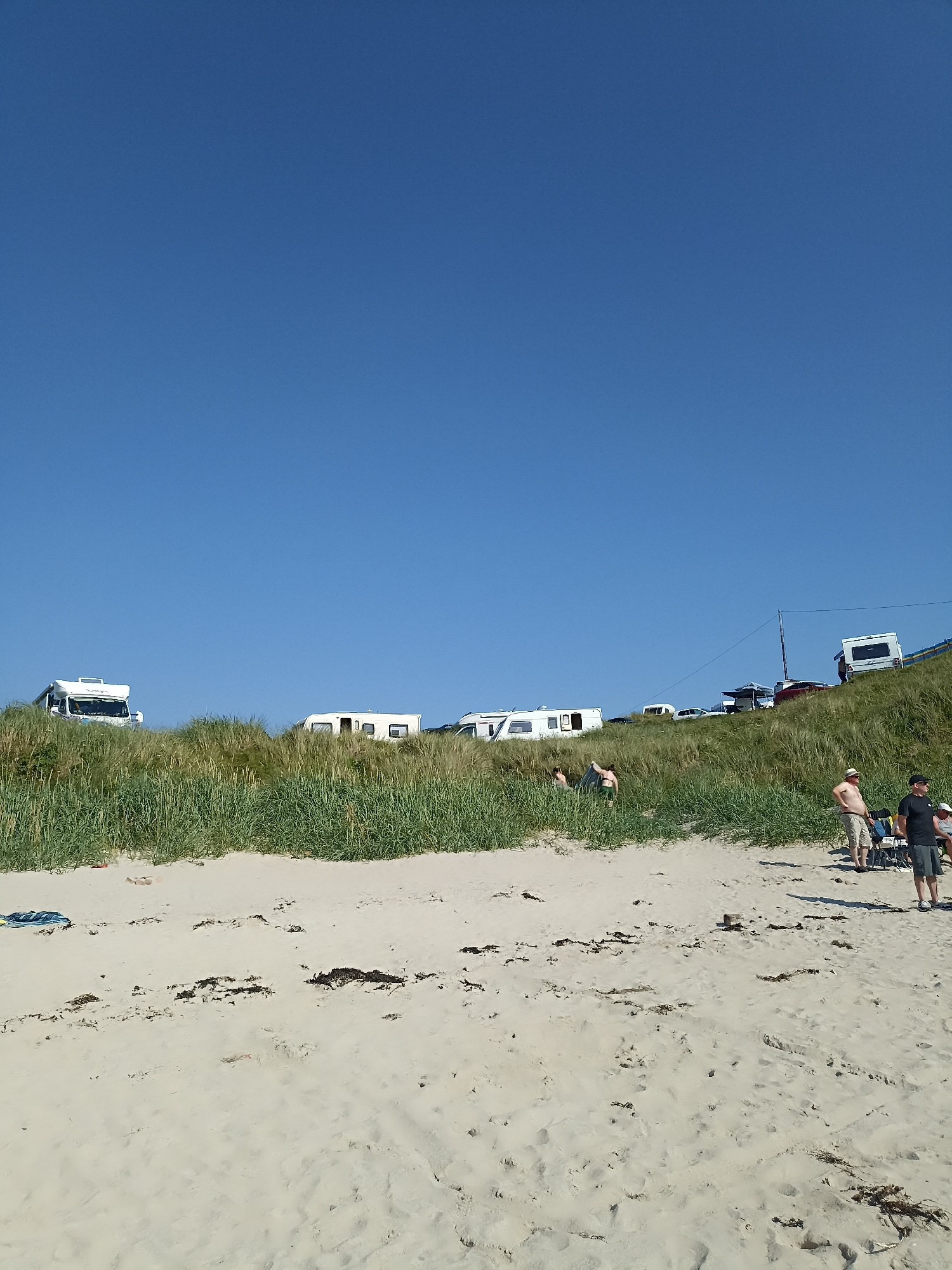 The image depicts a sandy beach with a clear blue sky. In the background, several white camper vans and a white caravan are parked on a grassy embankment. A few people are visible on the beach, some standing and others sitting. One person is wearing a hat, and two are wearing shorts. The beach is scattered with seaweed. There are also some chairs and a pole with wires in the scene. The location is identified as Carnboy, Ireland.