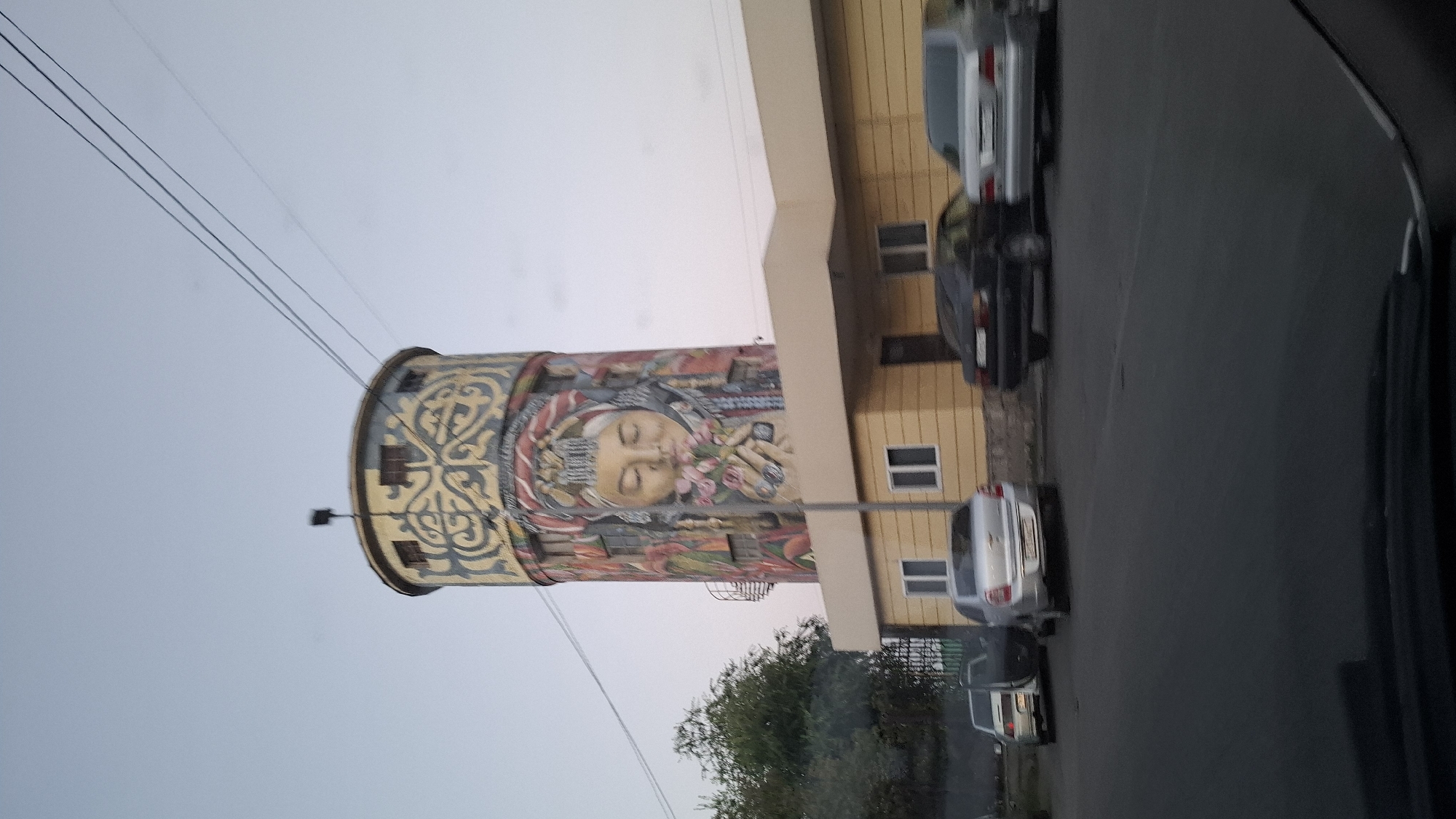 The image depicts a water tower in Shymkent, Kazakhstan, adorned with a large mural of a woman's face wearing traditional attire and holding flowers. The tower is cylindrical with decorative elements and windows near the top. Adjacent to the tower is a single-story yellow building with windows and a doorway, appearing to be a commercial or administrative structure. Several cars are parked along the street in front of the building and in the foreground. The environment suggests an urban setting with a clear sky.