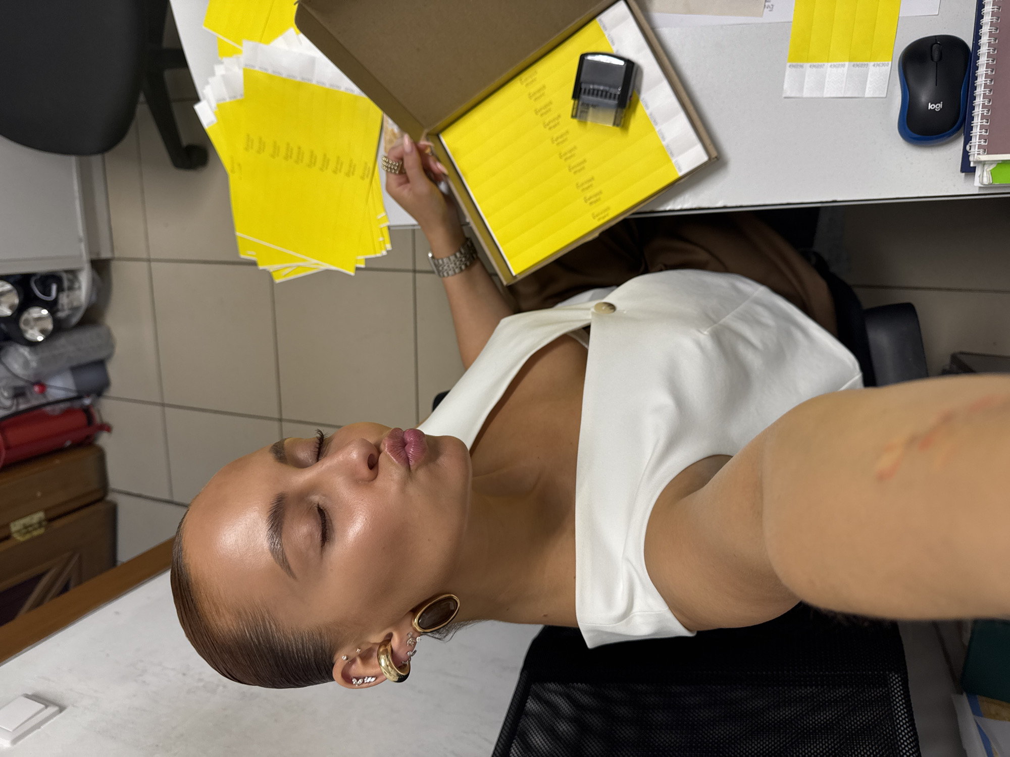 A forensic examination of the image reveals an indoor office setting, specified to be in the city of Chaikovsky, Russia. A female individual with medium complexion and slicked-back, reddish-brown hair is centered in the frame, taking a selfie. Her eyes are closed, and her lips are pursed in a kissing gesture. She is wearing a white sleeveless top with a deep V-neck and a button closure. Visible jewelry includes a gold helix cuff, multiple small stud earrings, and a large oval-shaped dark brown stone earring set in gold-toned metal on her left ear. A silver-toned watch is on her left wrist, and a ring with small stones is on her right hand.
The individual is seated at a light-colored desk, which occupies a significant portion of the lower and right frame. On the desk are multiple stacks of bright yellow paper strips, possibly wristbands or tickets, some loose and some contained within a brown cardboard box. A black self-inking stamp is positioned inside the box, adjacent to the yellow strips. A blue and black Logitech computer mouse is situated near the bottom right of the desk. Partially visible office supplies, including notebooks and binders, are also present on the desk.
To the left of the individual is a black office chair with a mesh back. The background consists of a light-colored wall with a white electrical outlet. A dark wooden door and frame are visible in the upper right background, along with indistinct metallic objects. The floor features light-colored square tiles. The lighting suggests an interior, possibly artificial light source.