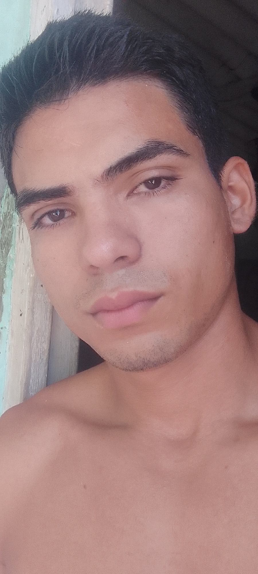 A close-up shot of a fair-skinned young man with dark hair and brown eyes. He has a short beard. The background shows a peeling wall with light green paint. He is shirtless, and the image appears to be taken outdoors. The location in Juan Griego, Venezuela is unknown.