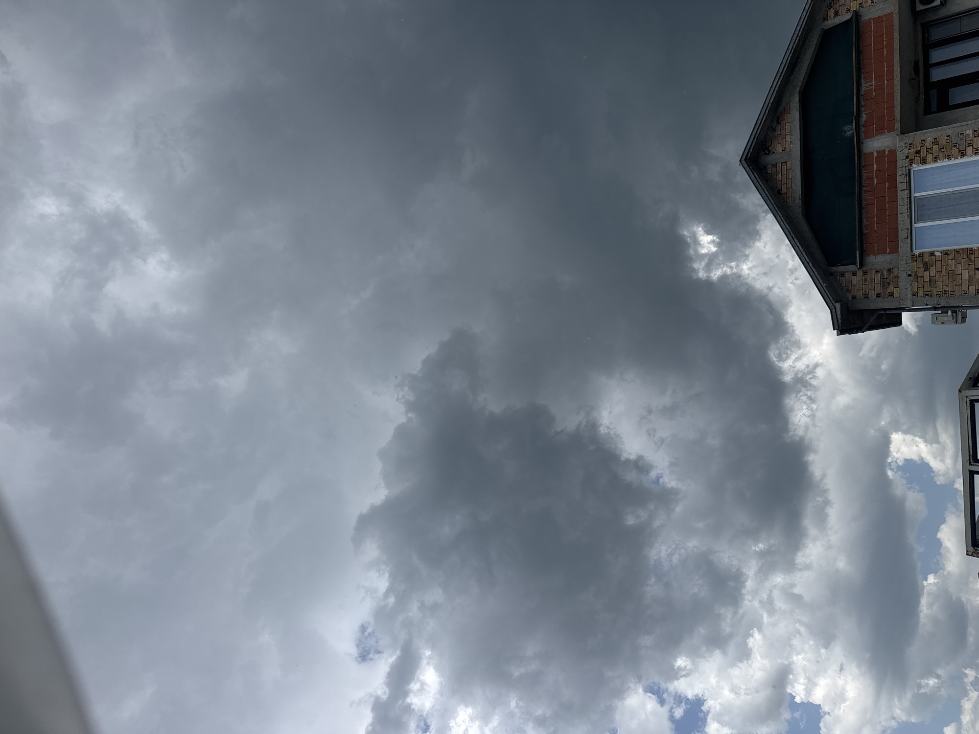 The image shows a view of a cloudy sky with a building partially visible in the upper right corner. The building appears to be under construction or renovation, with exposed brick and concrete. The sky is dominated by dark, ominous-looking clouds, suggesting an approaching storm. In the lower right corner, there are patches of blue sky visible. The building is constructed with brick and concrete blocks and partially finished with windows and a sloping roof. The image was taken looking upward. The exact location in the city of Sarnevo, Bulgaria, cannot be determined without further information or context.