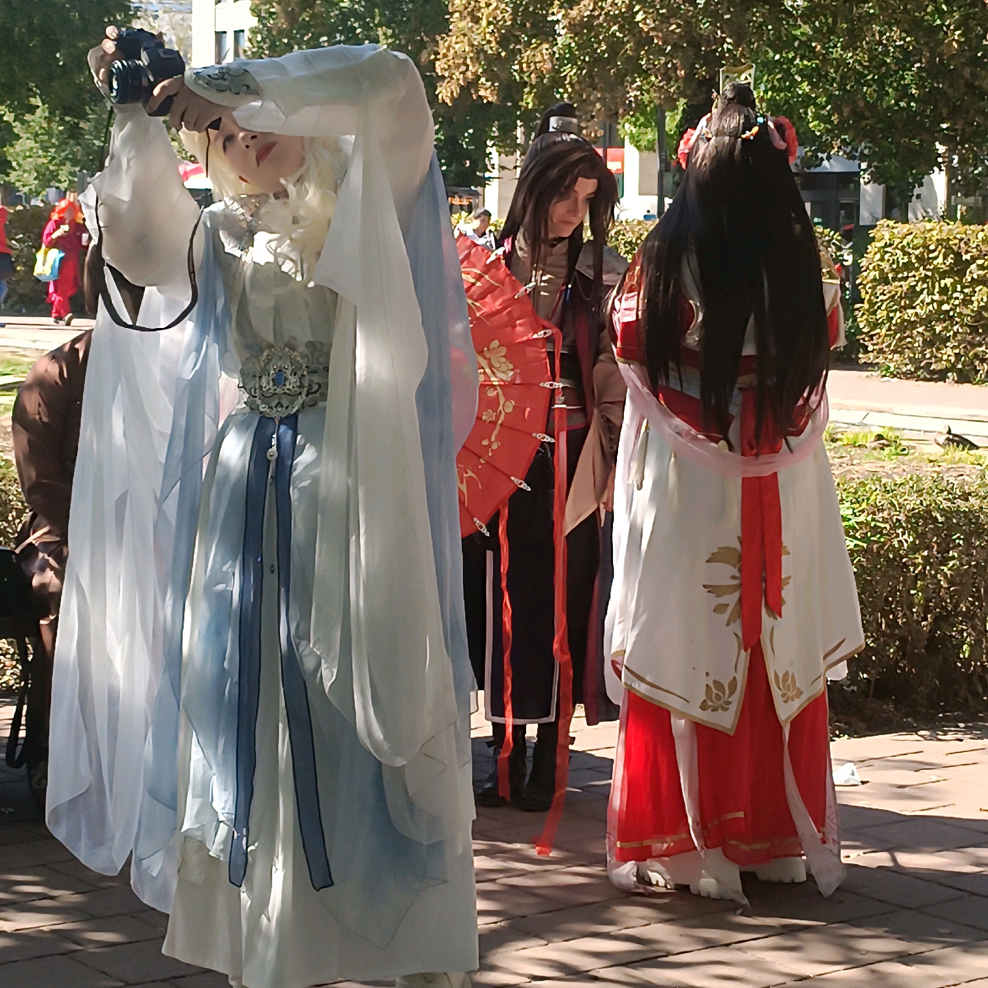 In the city of Wiesbaden, Germany, on an outdoor paved area, a group of individuals are posing for photographs. Two individuals in the foreground are dressed in elaborate costumes. The person on the left wears a white and light blue flowing robe with intricate silver detailing on the belt. They have light-colored hair and are holding a camera with their left hand, obscuring part of their face. The person on the right is dressed in a white robe with red accents, featuring gold floral patterns. They have long, dark hair adorned with pink accessories and are holding a red fan with a floral design. A third individual, partially visible behind the person on the left, wears dark clothing and also appears to be posing. The background shows trees with autumnal foliage, green bushes, and a building in the distance. The lighting suggests it is daytime, with shadows cast on the ground.