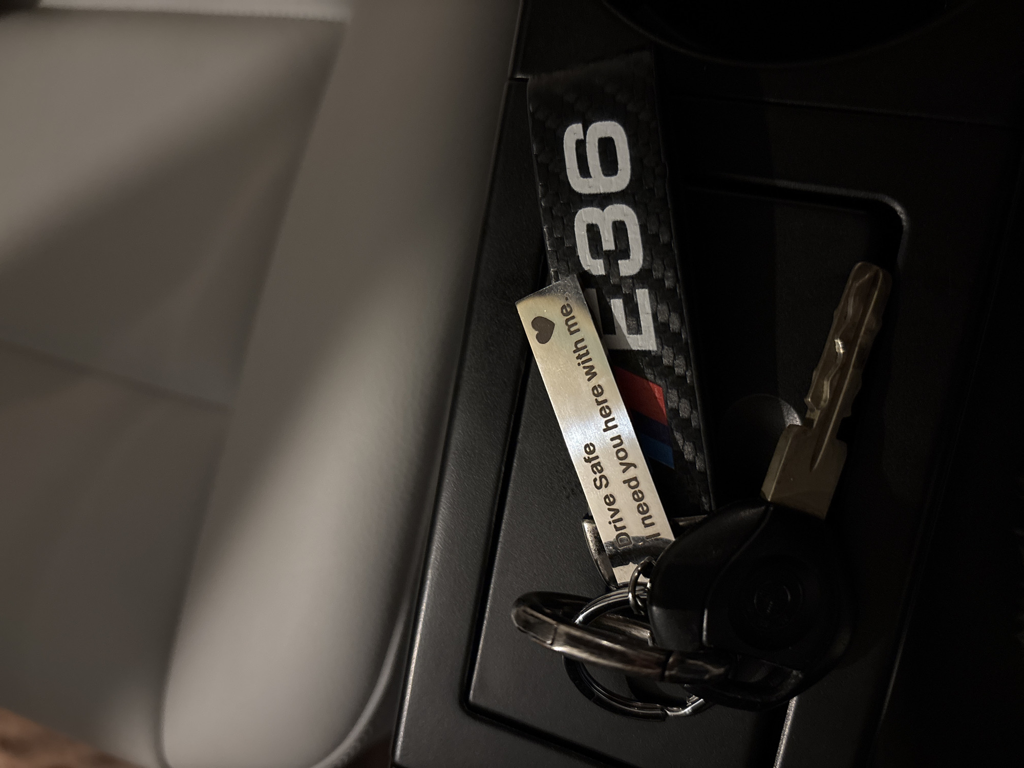 A set of car keys sits in the center console of a car. The keys are attached to two keychains. One keychain is black with "E36" printed on it in white. The second keychain is a silver rectangle with the words "Drive Safe. I need you here with me" printed on it next to a small heart. The car's seat is visible on the left of the frame and the rest of the console is visible on the right.