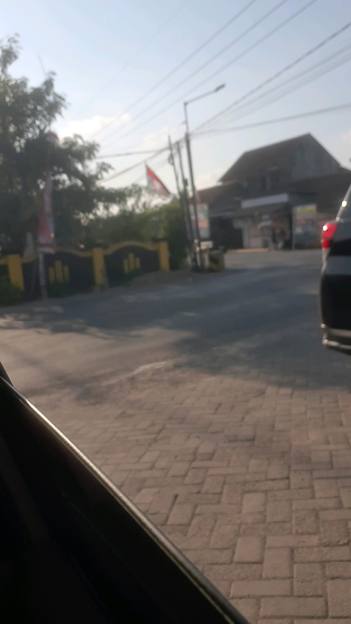 The image depicts a street scene in Seneporejo, Indonesia, with a focus on the foreground, which shows the edge of a dark vehicle and a paved walkway made of interlocking brick pavers. The walkway has a herringbone pattern. In the mid-ground, a paved road is visible, with a yellow and dark blue striped fence running along the side. Beyond the fence, there is greenery and a building with a dark roof. A flag, possibly red and white, is seen behind some trees. A street lamp is also visible, along with power lines overhead. The scene is captured during daylight, with the sun casting shadows. The image has a somewhat blurry quality, suggesting it might have been taken from a moving vehicle or with a camera that is not in sharp focus. There are no discernible people clearly visible or identifiable in the frame.