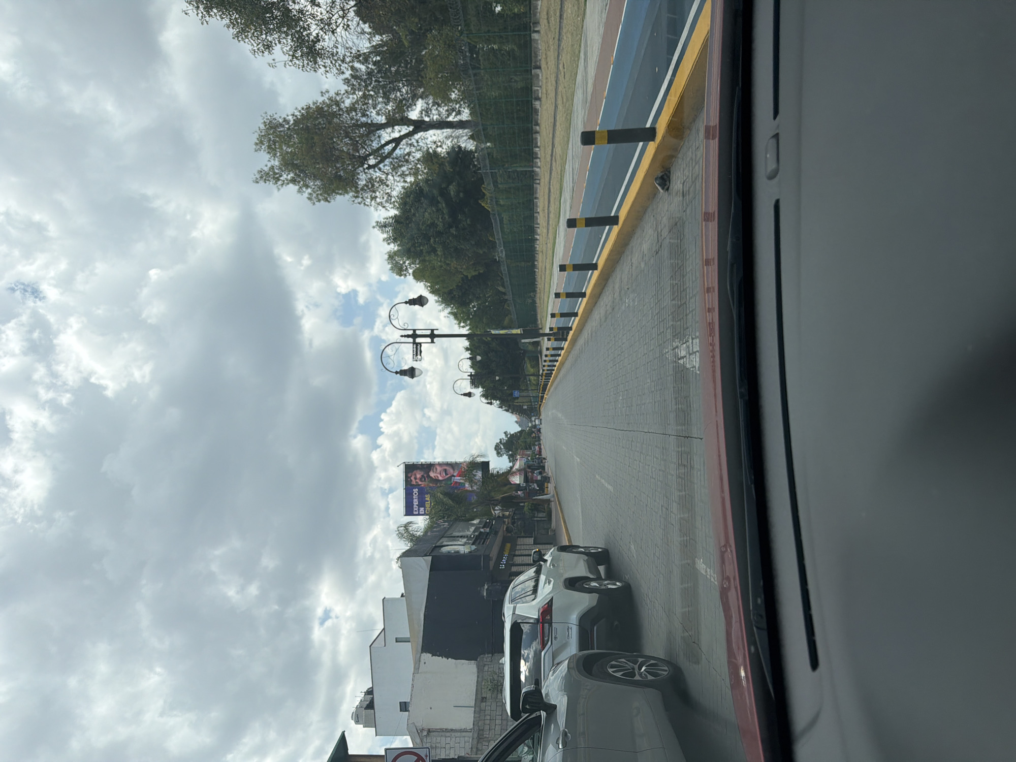 The image, taken from inside a vehicle, shows a street in San Andrés Cholula, Mexico. Traffic is moderately heavy with several cars visible. The road is paved with light gray tiles and features a pedestrian walkway with a yellow curb. A large billboard displaying faces and Spanish text is visible on the left side of the street. Street lights are also present. The sky is overcast with white and gray clouds. Trees line the sidewalk. A portion of the car's interior is visible in the foreground.