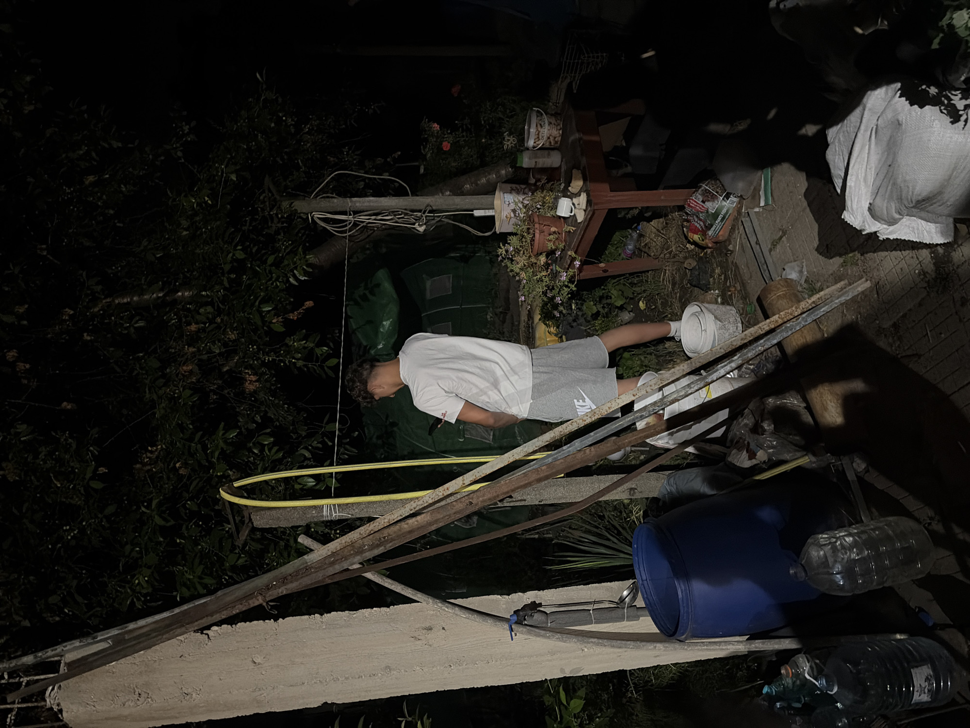 In the city of Novi Iskar, Bulgaria, a young man stands outdoors at night, dressed in a white t-shirt and gray shorts. He is holding a mobile phone in his right hand and appears to be urinating. The surrounding environment is cluttered, with various objects visible, including a large blue barrel, plastic water bottles, a wooden table with potted plants and containers, and a green structure in the background. The scene is dimly lit, with shadows obscuring some details. The ground is uneven and covered with debris.