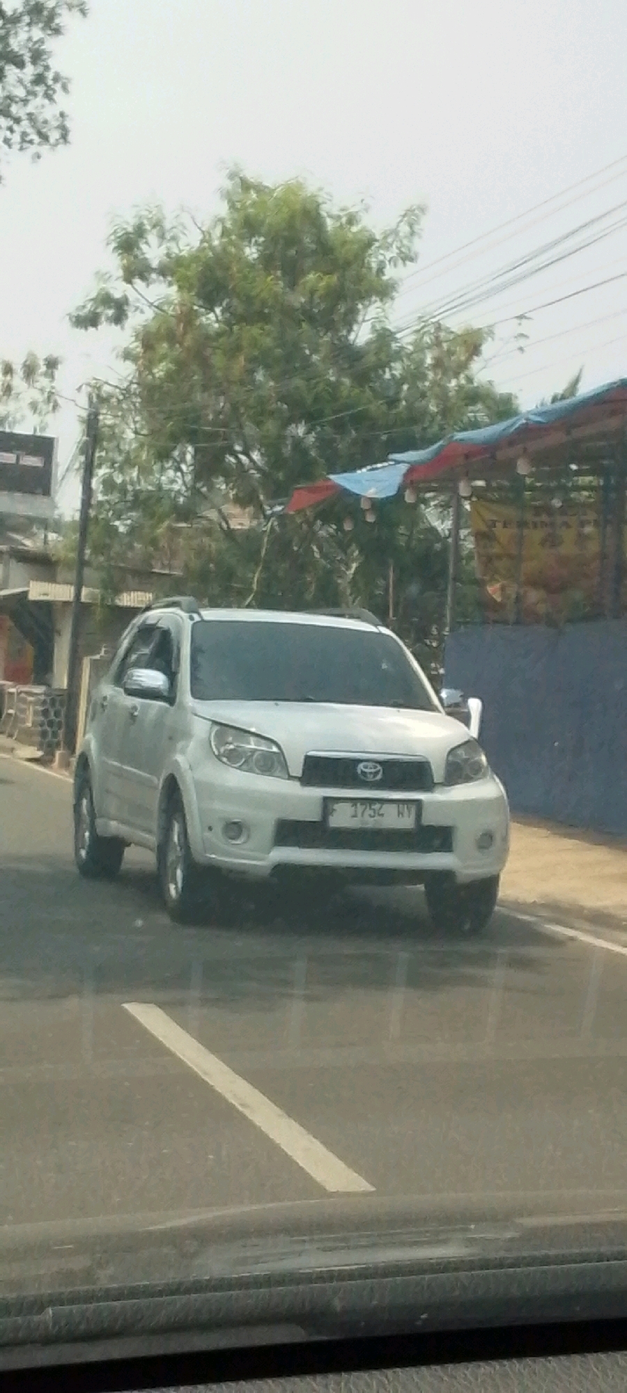 A white Toyota Rush is parked on the side of a road in Cisalak, Indonesia. The vehicle is facing forward, and the license plate is visible and reads "F 1752 WV." In the background, there are trees and buildings, including a structure with a blue and red awning. The sky is overcast. The image appears to have been taken from another vehicle.