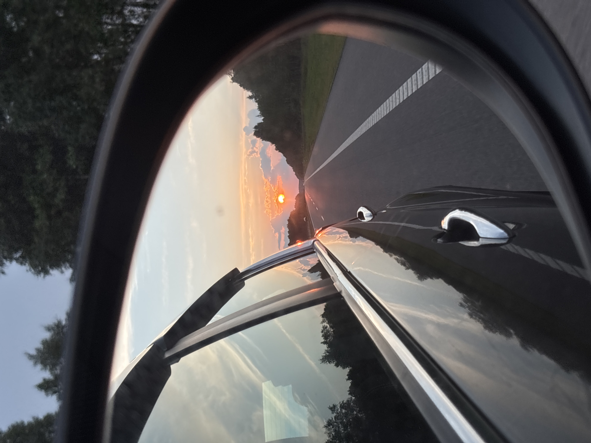 The image is a rearview mirror reflection showing a sunset over a highway. The reflection captures the sky with scattered clouds and the sun partially obscured by clouds, casting a warm orange glow. A dark-colored car is visible in the foreground, reflecting the sky and surrounding trees. The highway stretches into the distance, with clear lane markings. Dense trees line the right side of the highway. The overall scene suggests travel on a road in a rural or suburban area during twilight. There are no people visible in the image. The location is identified as Yassyenki, Belarus, which is a village in the Yassyenki rural council of the Mogilev Region in Belarus.