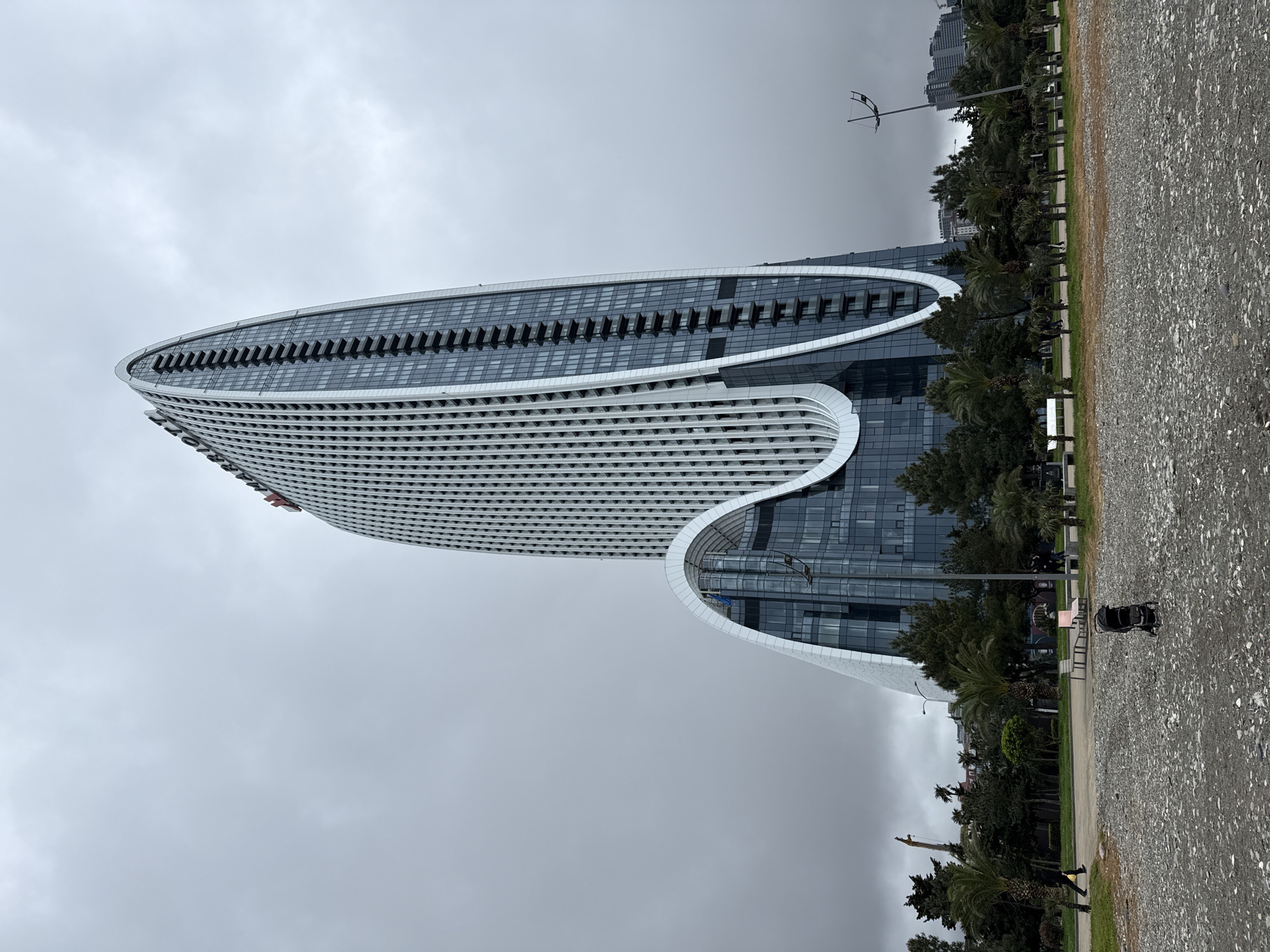 The photo shows a tall, modern building in Batumi, Georgia. The building has a unique, wave-like design with a white facade and many windows. A row of trees is on the right side of the building, with the ocean behind it. The sky is cloudy and gray. A paved path runs along the right side of the building, and a stroller is parked near the trees. A street lamp is visible in the distance.