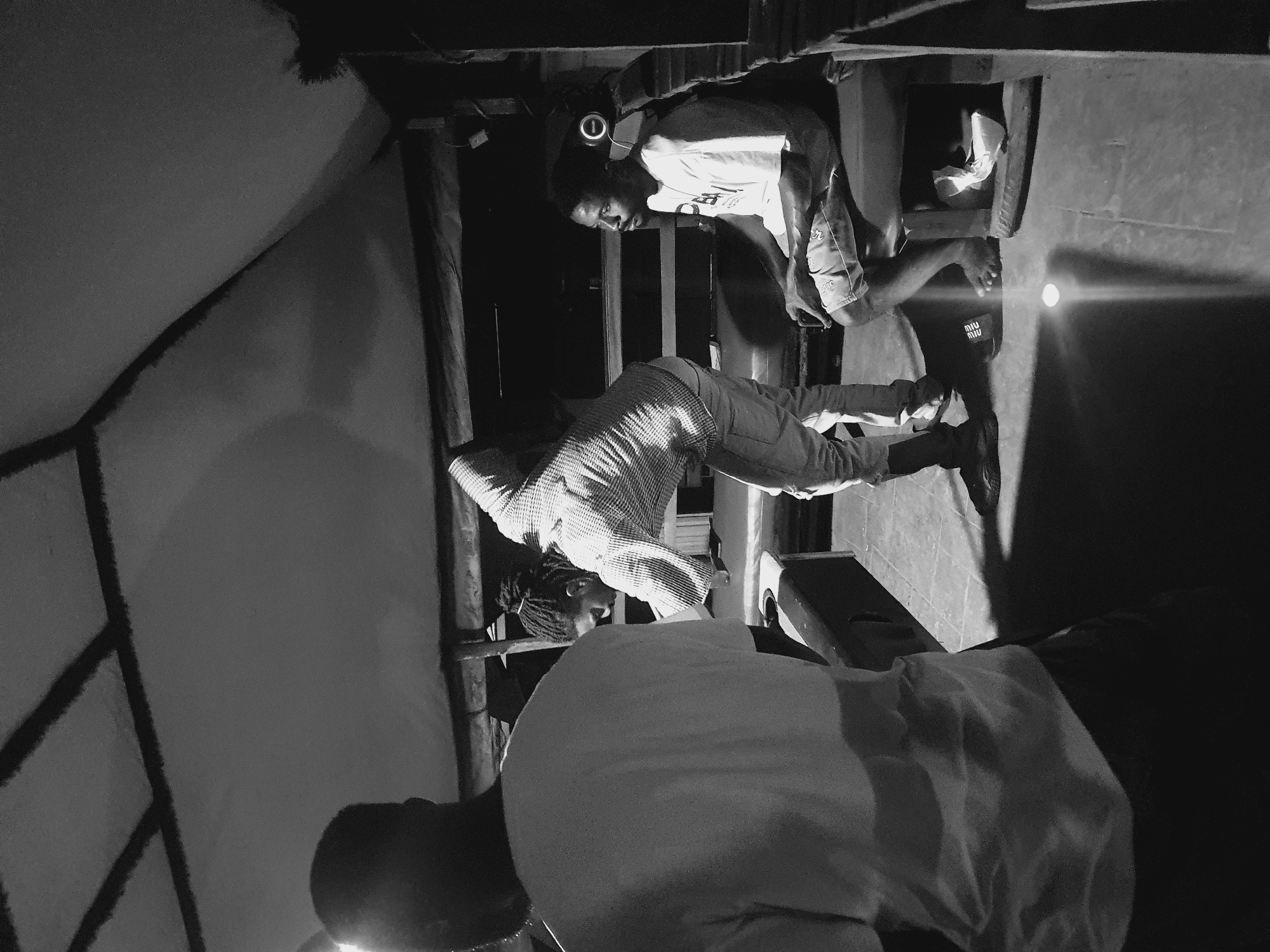 The black and white image depicts an indoor scene with three individuals. In the foreground, on the left, a person stands with their back to the camera, wearing a light-colored t-shirt and dark trousers. In the middle ground, a person with dreadlocks, wearing a checkered shirt and light-colored trousers, is leaning over a surface, possibly a table or counter, and appears to be engaged in an activity. To the right, a man is seated on a bench, wearing a t-shirt and shorts, with headphones around his neck. He is holding a dark object in his hands. The environment appears to be a covered outdoor space, with a textured ceiling supported by beams, suggestive of a pavilion or open-air structure. The lighting is dim, casting shadows and creating a moody atmosphere. The scene does not provide any direct visual cues to ascertain the specific location in Lokoja, Nigeria.