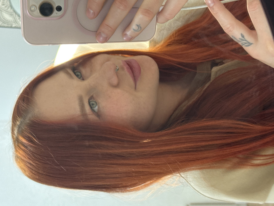 A selfie features a young woman with striking red hair, freckled skin, and light eyes. She has a nose ring and two small tattoos on her fingers. She's holding a pink phone with visible camera lenses. The angle is slightly rotated, showcasing her face from a unique perspective. Part of her body is visible in light-colored clothing. The background appears to be a light, neutral color.