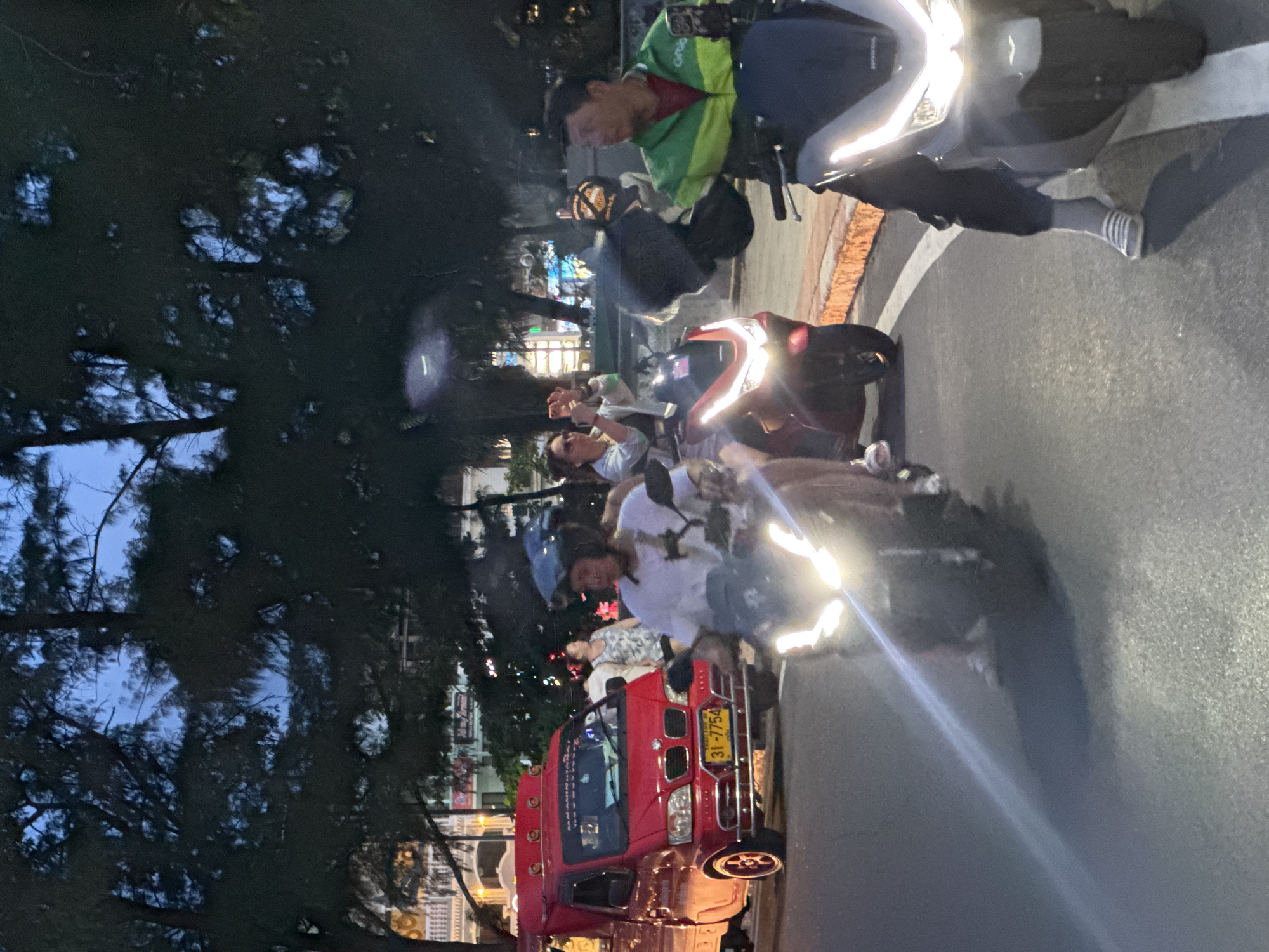 The image depicts a street scene in Karon, Thailand, likely during the evening or dusk. In the foreground, two individuals are riding motorcycles. The motorcycle on the right is a dark color with bright headlights, and the rider is wearing a green and black jacket with "Grab" branding, suggesting they are a ride-sharing driver. They are holding a dark helmet. The motorcycle on the left is a lighter color and its rider is also wearing a helmet. In the background, a red pickup truck, possibly a songthaew, with a license plate reading "31-7754" is visible. Other pedestrians and buildings can be seen in the distance, with some lights illuminating the scene. Trees with dense foliage line the street, and the sky is a deep blue, indicating it is not yet fully dark. The road surface appears to be asphalt with white lane markings.