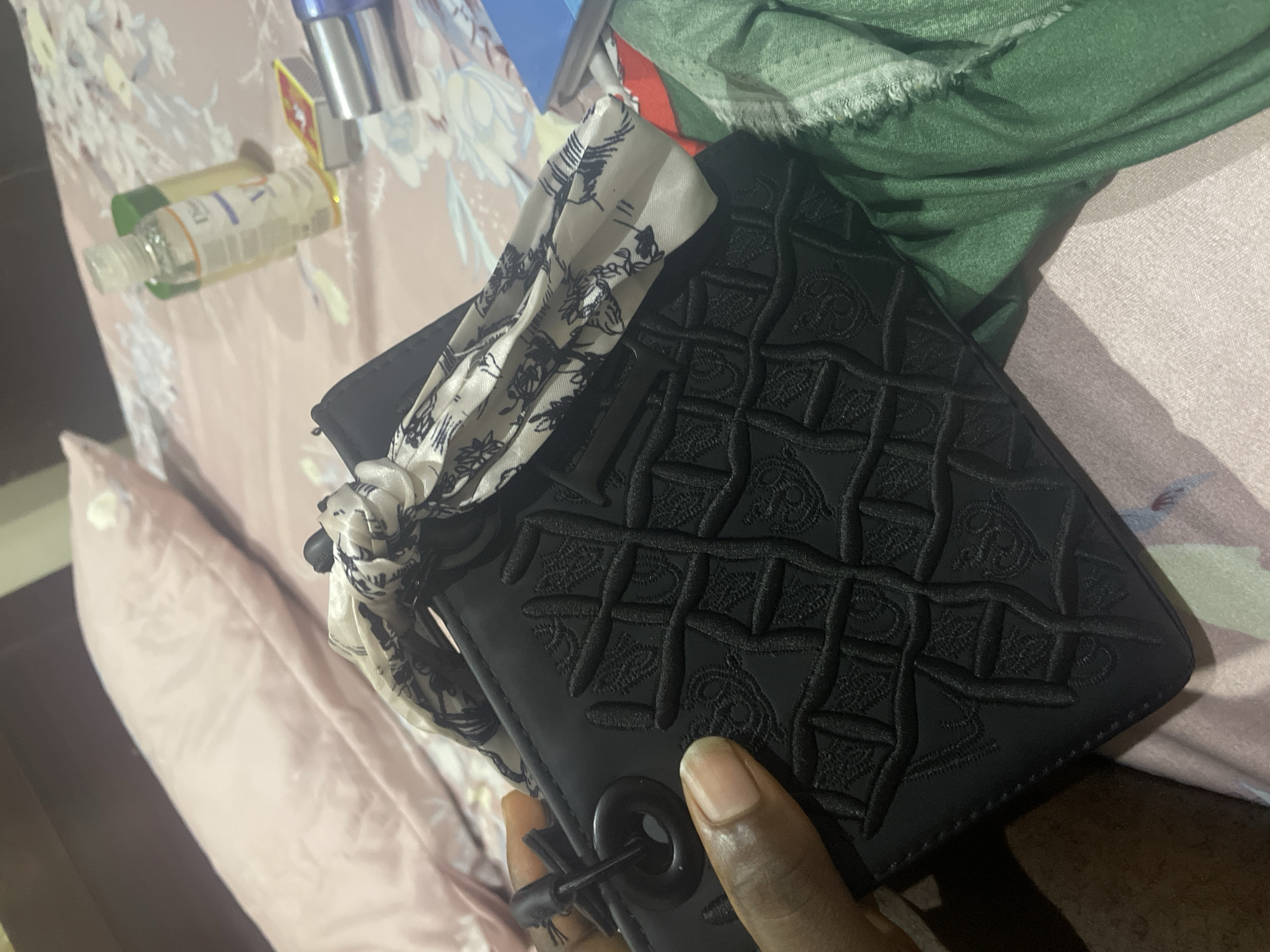 A person's hand is holding a black handbag with a patterned design and a scarf tied to its handle. The handbag is resting on a bed with pink pillows and a floral-patterned bedsheet. In the background, there are bottles of hand sanitizer and a box of matches. The scene appears to be indoors, likely in a bedroom in Jalingo, Nigeria.