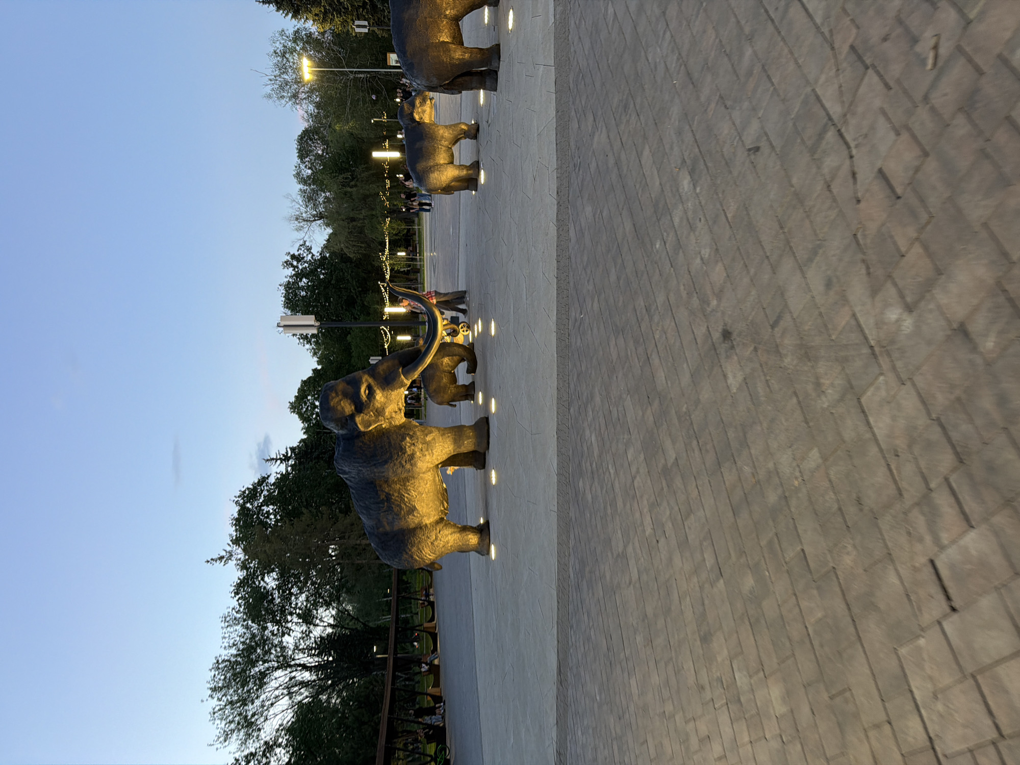 In the city of Unknown, Unknown, at dusk, several large, dark-colored sculptures of mammoths are displayed on a paved area. The sculptures are illuminated by spotlights, casting a warm glow on their textured surfaces. The largest mammoth sculpture is in the foreground, facing right, with its trunk curved. Two smaller mammoth sculptures are positioned behind it, and another is visible to the far right. The background features trees, lampposts, and several indistinct figures of people walking. The sky is a gradient of pale blue and purple, indicating the transition from day to night. The ground is paved with rectangular stones in varying shades of gray and brown. In the mid-ground, a wooden structure with suspended seats is visible, with a few people seated on them. The scene appears to be an outdoor public space, possibly a park or plaza, designed for aesthetic display and public enjoyment.