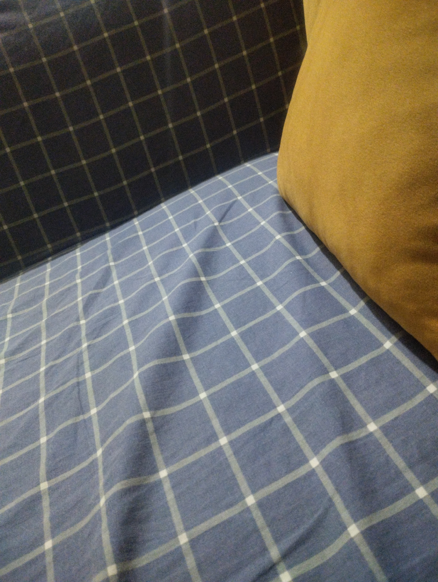 A close-up, overhead shot shows a brown pillow resting on a blue and white checkered fabric. The fabric is wrinkled and appears to be a bedsheet or a similar material. The pillow is soft and appears to be made of suede or a similar textured fabric. The background is a dark blue fabric with a lighter blue checkered pattern. The scene is lit by a warm, soft light, suggesting an indoor setting, possibly a bedroom. The mood is calm and cozy. There is no visible text in the image.