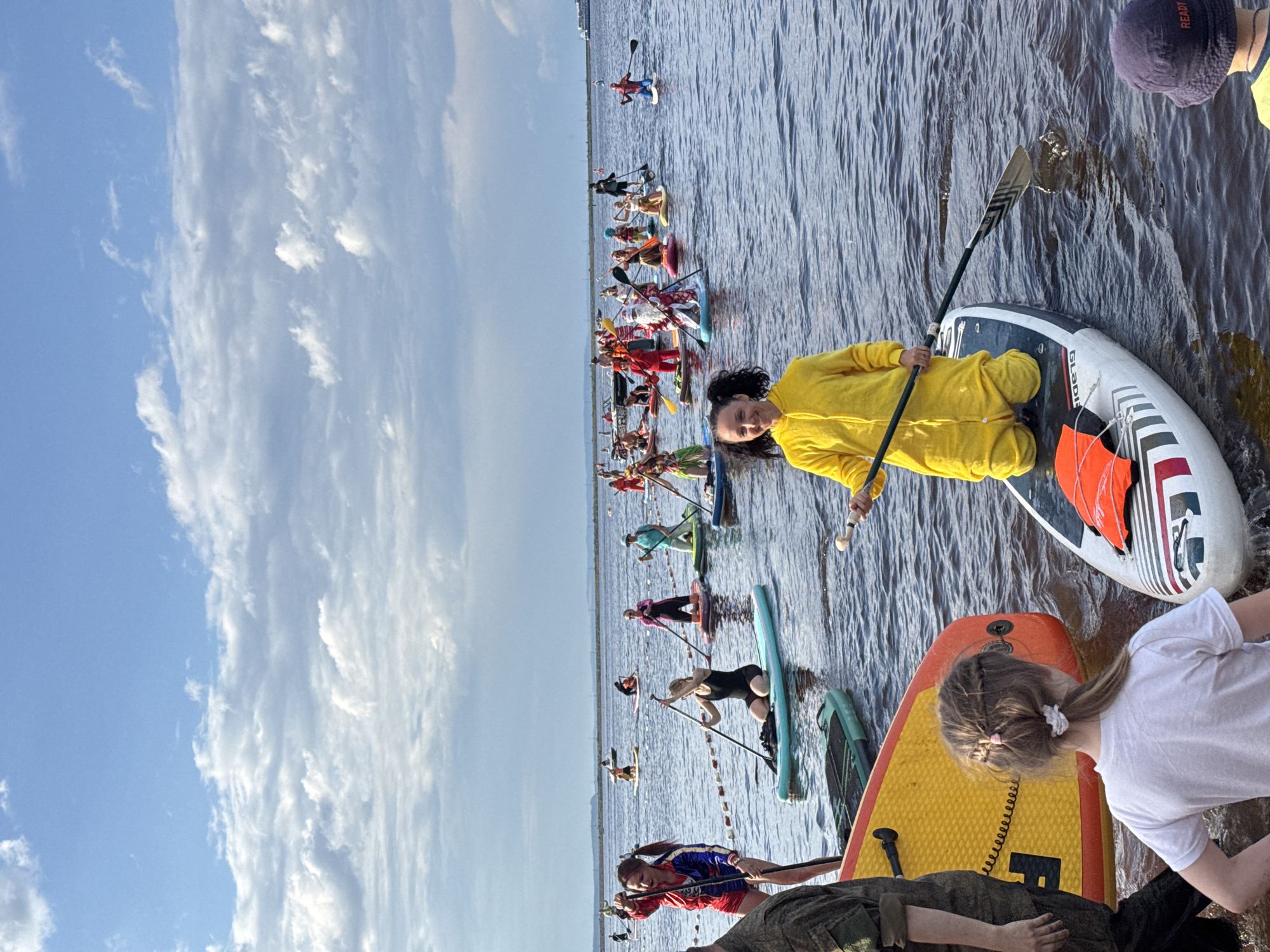 This is a wide shot of a large group of people participating in a paddleboard event on a body of water, likely during daytime given the bright sky. The sky is partly cloudy, with large, fluffy white clouds against a blue background. The water is dark and appears to be relatively calm with some ripples.
In the foreground, on the right, a person wearing a yellow hooded jumpsuit is kneeling on a paddleboard, holding a paddle. To the left of this person, a young girl with braided hair tied back is wearing a white t-shirt. Further to the left, a person in a camouflage uniform is visible from the waist down. In the mid-ground, there are numerous other individuals on paddleboards. Some are paddling, while others are stationary. Many are dressed in costumes, including what appears to be a Santa Claus costume, a Spider-Man costume, and others in brightly colored outfits. There are also buoys or markers in the water, creating a line that separates the participants.
In the background, more people on paddleboards are visible, extending into the distance. A ferry or large boat is present on the horizon. The distant shoreline is lined with trees or vegetation, and the sky continues with clouds. The overall impression is of a festive event or competition taking place on the water. The location is identified as the Kama River in the city of Chaikovsky, Russia.