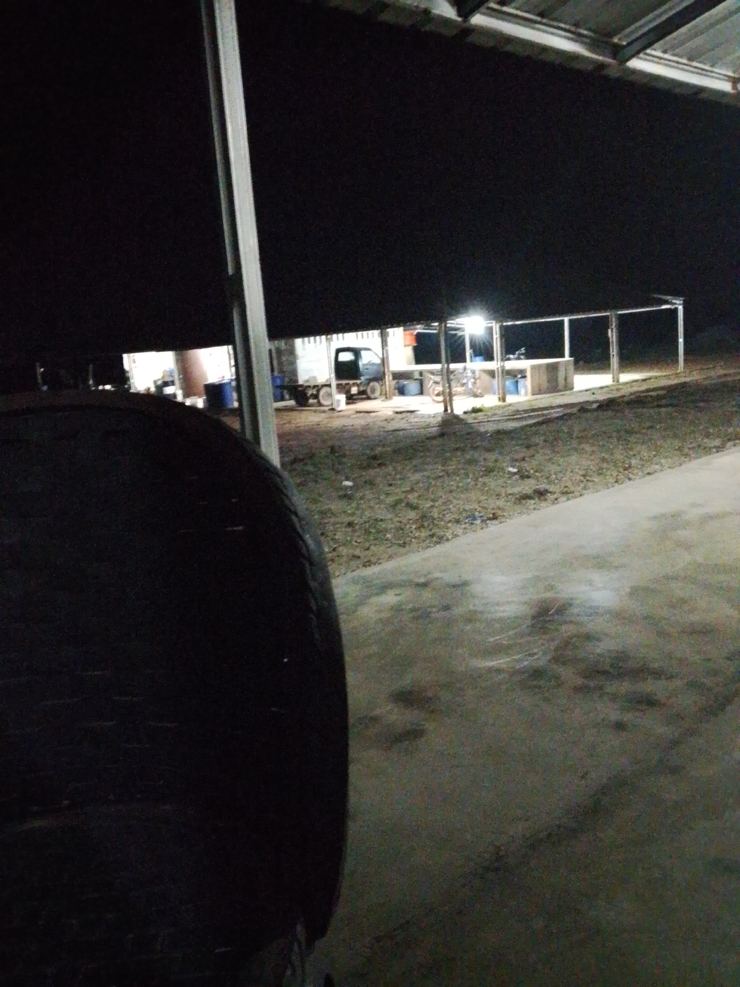 The image was taken at night in Bukit Ketok, Indonesia, under a partially covered structure. A large, dark tire is prominent in the foreground, occupying the left side of the frame. Beyond the tire and to the right, a concrete or paved surface extends into the middle ground. Farther in the background, several open-sided structures are visible under artificial lighting. A small truck is parked near one of the structures. Debris and ground cover surround these structures. The sky is dark, suggesting nighttime. The image has limited lighting and lacks human presence.