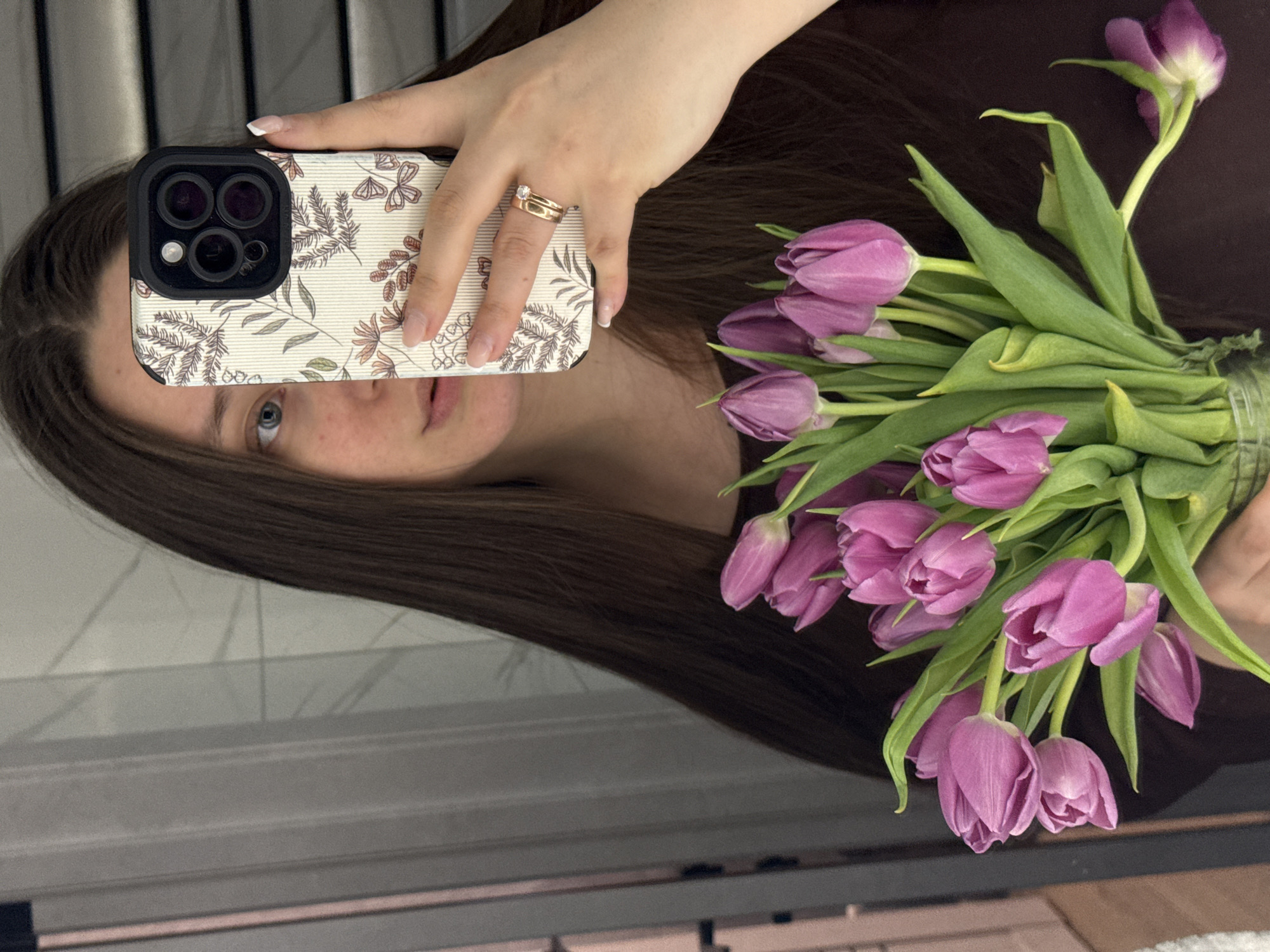 A female subject is depicted in a mirror selfie, partially obscuring her face with a smartphone held in her right hand. The smartphone, likely an iPhone due to its camera array, features a light-colored case adorned with botanical and butterfly patterns in brown and pink hues. On the subject's right ring finger, two gold rings are visible: a plain band and one set with a single clear stone. Her fingernails exhibit a light-colored polish with white tips. Her left hand holds a bouquet of approximately 15-20 closed purple tulips with green foliage. One tulip stem appears detached or significantly drooped at the lower right portion of the bouquet. The subject has long, dark brown hair and light-colored eyes; a portion of her left eye and cheek with freckles are visible. She is wearing a dark, solid-colored garment. The background, visible as a reflection, consists of indistinct light grey vertical elements, possibly window coverings or wall paneling. Specific location details within Krasnodar, Russia, are not identifiable from the visual content.