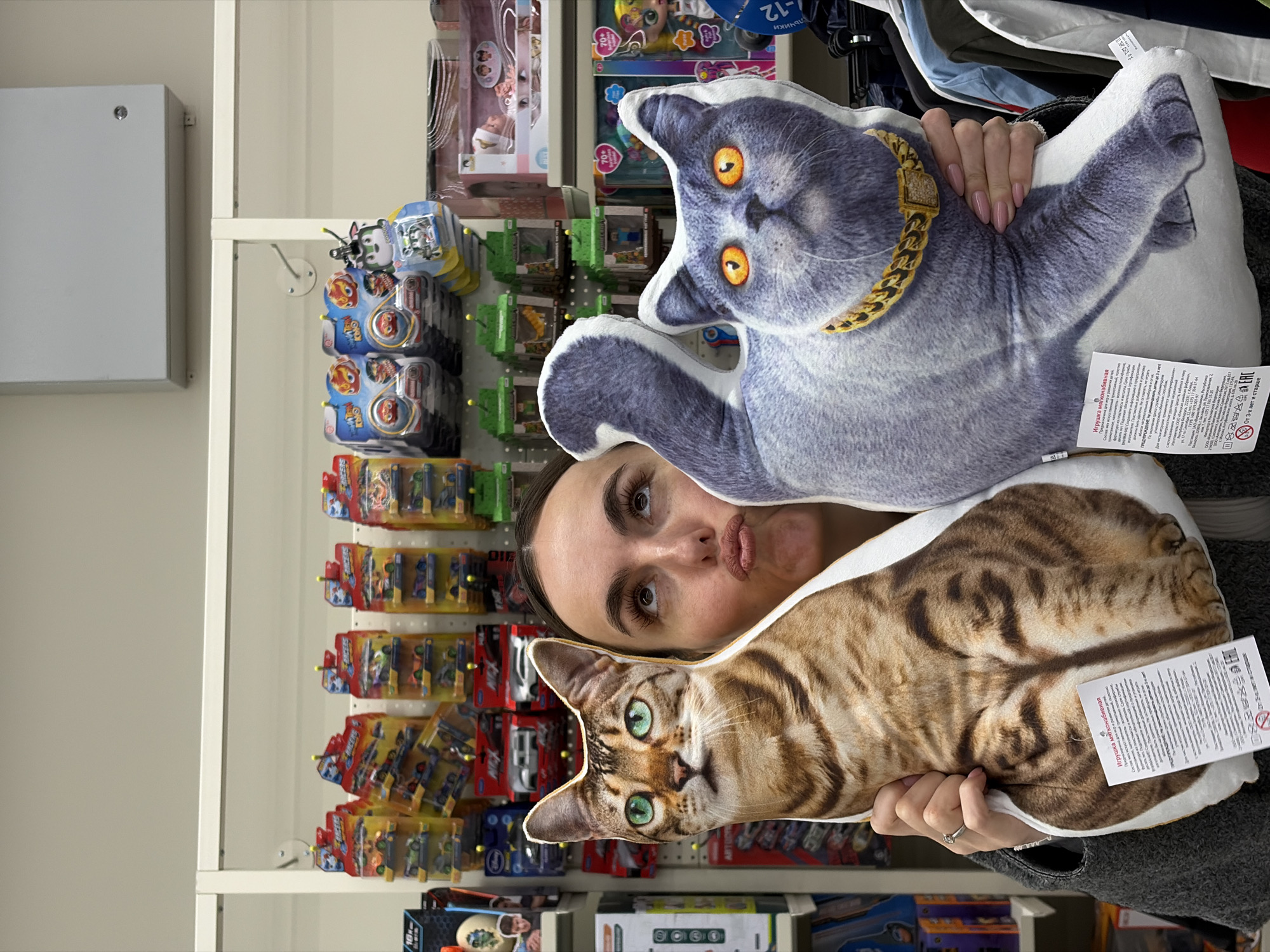A woman with dark hair and a neutral expression is holding two cat-shaped pillows in front of her face. One pillow features a grey cat wearing a gold chain, and the other depicts a brown tabby cat. Behind the woman and the pillows are shelves stocked with toys, including boxed action figures and dolls. The environment appears to be a store, specifically a toy section. The tags on the pillows include Russian text. The OCR text confirms the location: Izhevsk, Russia.