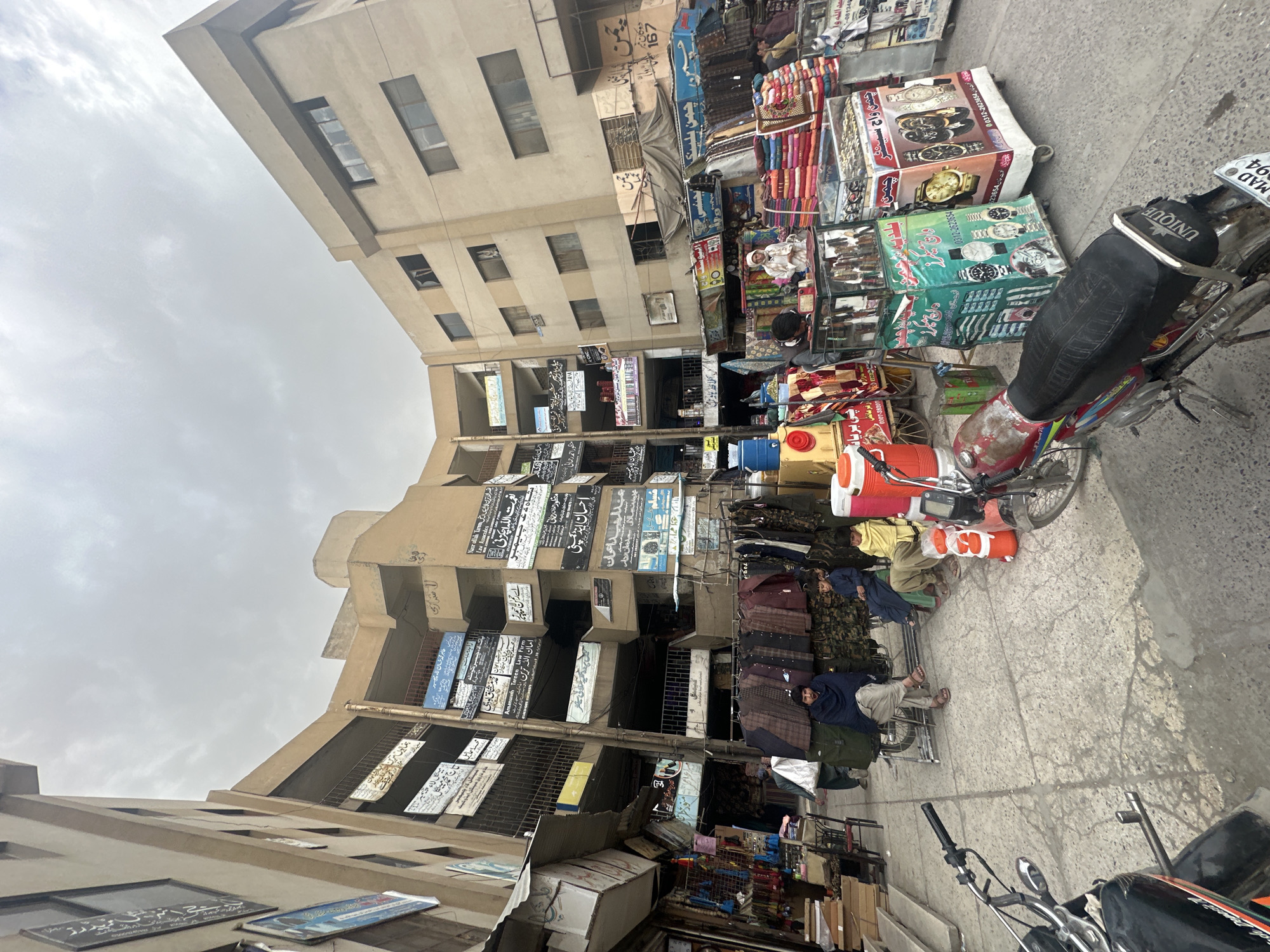 The image depicts a street scene in Quetta City Tehsil, Pakistan, with a narrow sidewalk lined with vendors selling various goods. The items for sale include clothing, watches, and other miscellaneous merchandise. Several people are present, either selling or observing the goods. Two motorcycles are parked on the sidewalk. A tall building forms the backdrop of the scene, its facade adorned with numerous signs written in Urdu. The sky overhead is overcast.