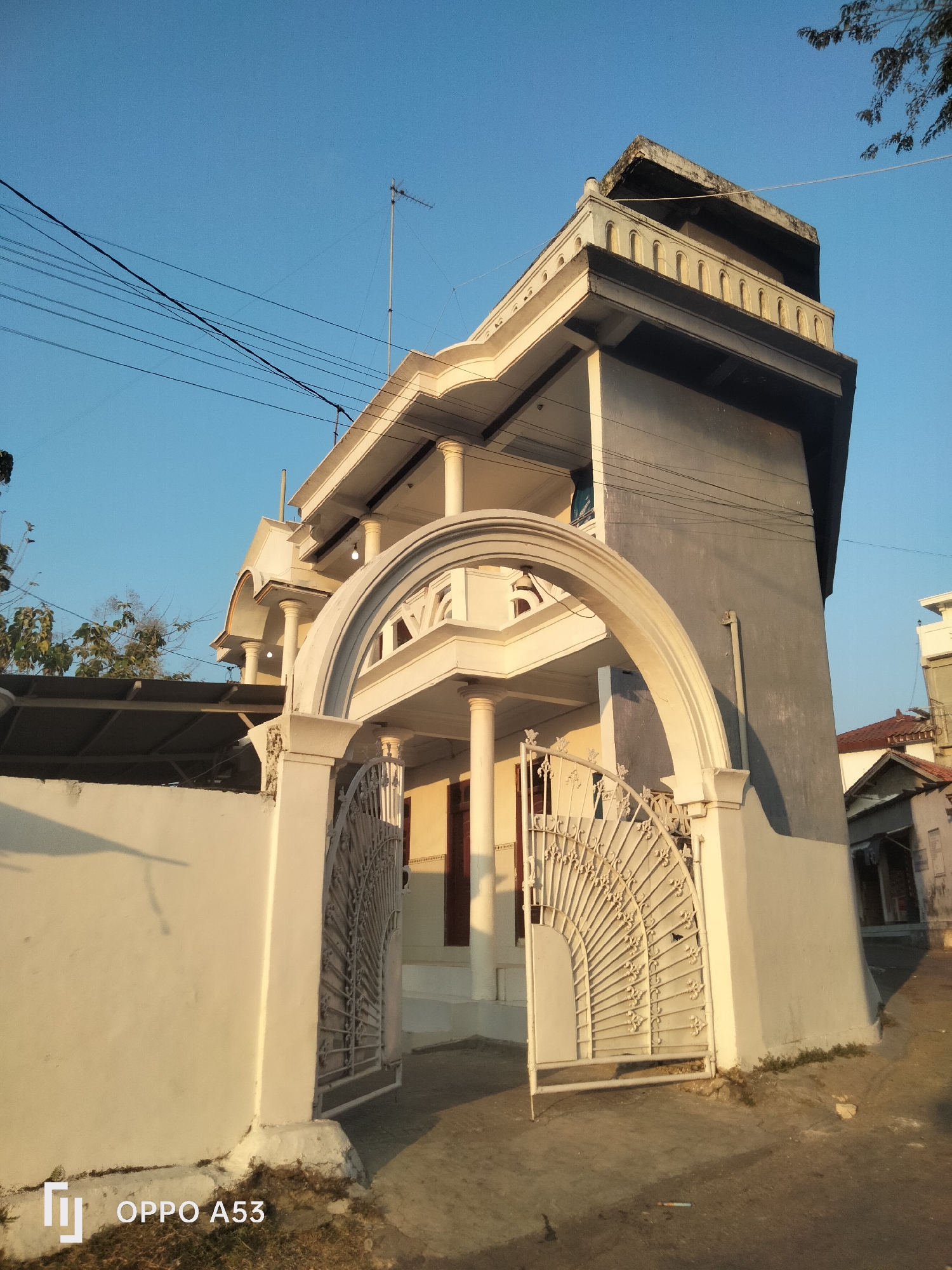 This is a full-frame, eye-level, outdoor, daylight, color photograph of a two-story building in Badung, Indonesia. The building has a gray facade and white architectural details including a large arched entryway supported by columns and a decorative white metal gate that is partially open. The building's upper story features a balcony with a decorative railing. Overhead, power lines crisscross the clear blue sky, and a television antenna is visible on the roof. To the left of the building, a white wall encloses a property, and some trees with green foliage are visible. The ground in front of the building is paved with a rough surface, and there is a patch of dry grass to the lower right. A watermark with the OPPO A53 logo is present in the lower left corner of the image.