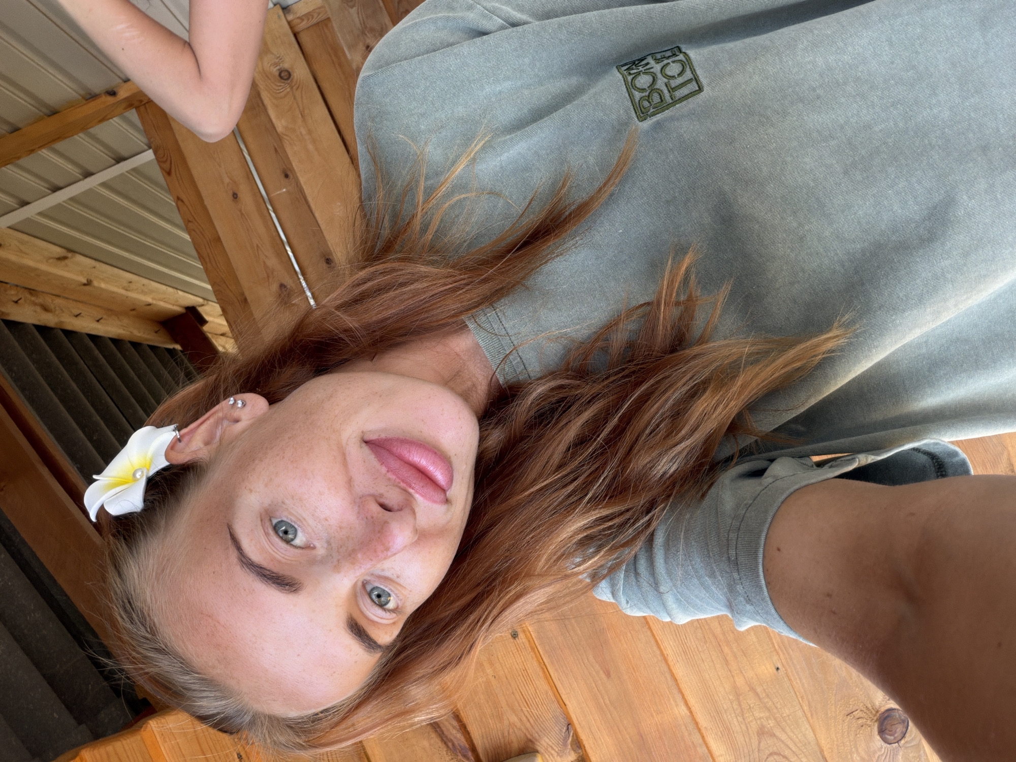 A woman with auburn hair, blue eyes, and freckles is taking a selfie. She is wearing a light green t-shirt with a small square logo on the left chest that says "BORN TO DIE". A white plumeria flower with a pearl is tucked behind her right ear, and she has a silver earring in her right earlobe. She is outdoors, with wooden structures visible in the background, suggesting she might be on a porch or deck. The lighting indicates it is daytime.