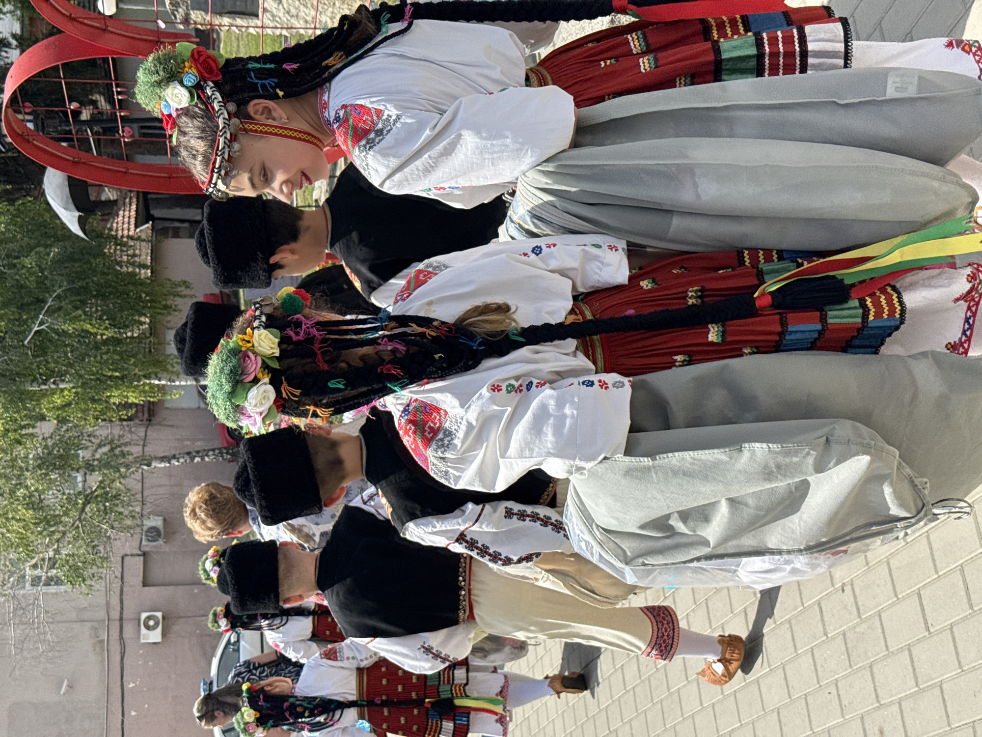 A group of children, dressed in traditional Bulgarian attire, are walking down a paved street in the city of Pordim, Bulgaria. The children are adorned with colorful embroidered shirts, vests, and skirts, as well as floral headpieces. They are wearing black hats and appear to be participating in a parade or festival. The background shows buildings, trees, and a red archway, suggesting a public event is taking place. The lighting indicates it is a sunny day.