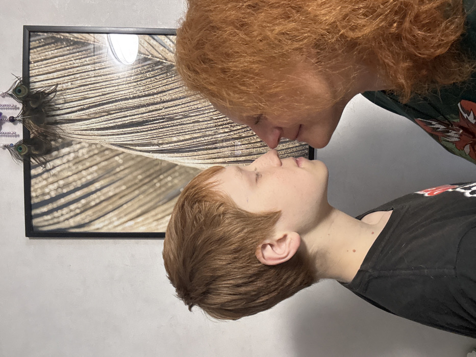 The image depicts an indoor scene featuring two individuals, a younger person and an older person, both with prominent reddish-orange hair, engaging in a nose-to-nose interaction.
The individual on the left appears to be a male-presenting youth with short, textured reddish-brown hair cut close on the sides and slightly longer on top. He has a pale complexion, a slightly upturned nose, and his eyes are closed. He is wearing a dark-colored t-shirt, possibly black, with a faint graphic partially visible on the upper left chest area.
The individual on the right is an adult female-presenting person with abundant, curly reddish-orange hair. Her profile shows a rounded nose and her eyes are closed, similar to the younger individual. She is wearing a dark garment, possibly a t-shirt or blouse, with a visible colorful pattern or design featuring red and green elements on her right shoulder area.
Their noses are lightly touching, and their eyes are closed, suggesting a moment of affection or connection.
The background consists of a light-colored, possibly off-white or light gray, wall. Centered behind the two individuals is a rectangular framed picture with a black border. The artwork within the frame features a textured, abstract pattern predominantly in shades of gold, brown, and black, resembling natural fibers, feathers, or plant structures. Above the framed picture, near the top edge, is a small decorative item comprised of what appear to be peacock feathers and dark-colored beads. A reflection from an overhead light source is visible on the upper right side of the framed picture.
The scene takes place indoors, likely within a private residence in the city of Chaikovsky, Russia. No further specific location details within the city are discernible from the image.