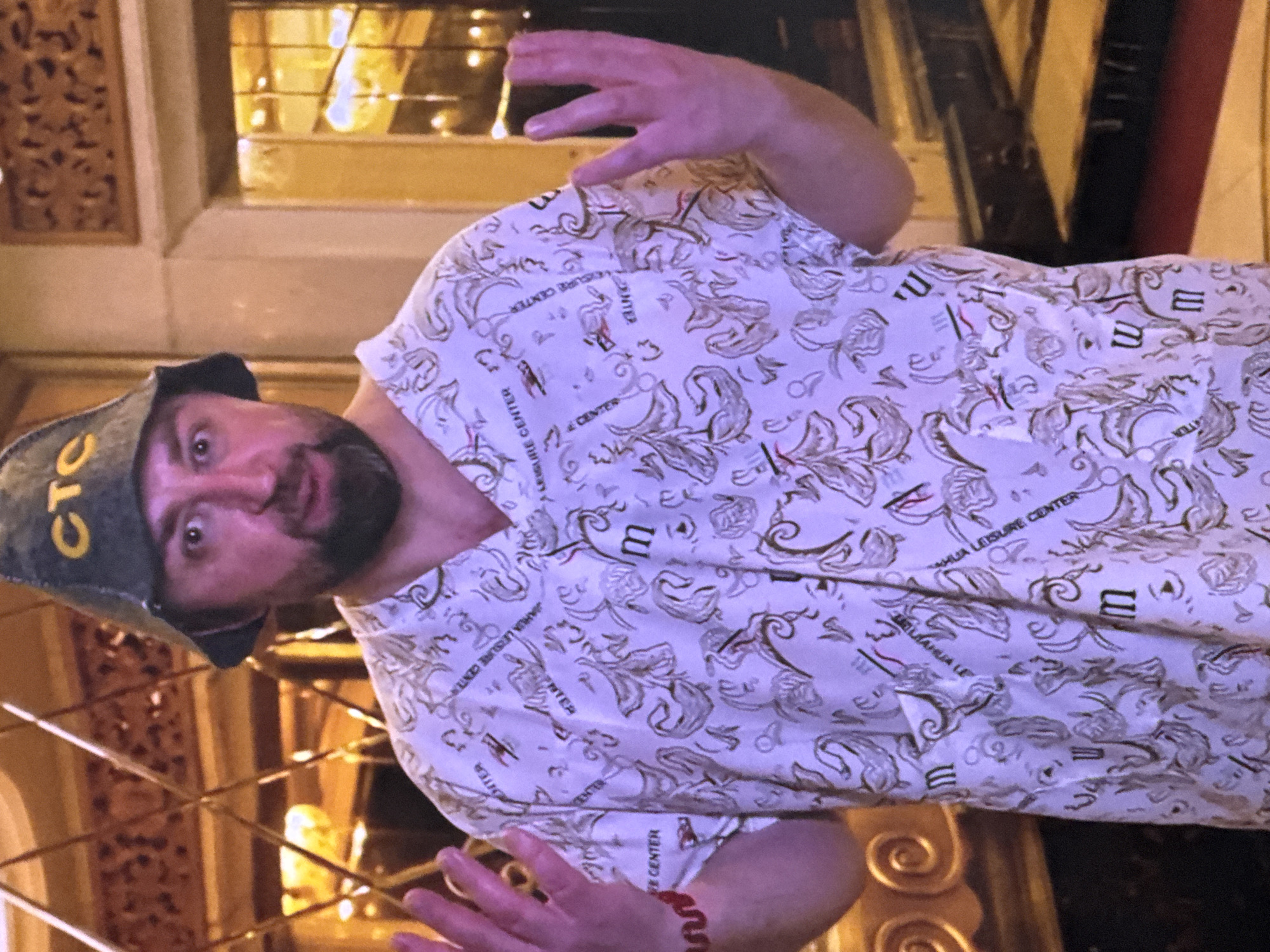 A man with a beard and a dark green hat with yellow lettering that reads "CTC" is standing in what appears to be a hotel lobby. He is wearing a white short-sleeved medical scrub top with a repeating pattern of leaves and some text that appears to read "AMHUA LEISURE CENTER". He has his hands raised, palms facing outwards, and is looking directly at the camera with a slightly surprised expression. A red beaded bracelet is visible on his left wrist. The background shows ornate gold and brown paneling and lighting that suggests a luxurious indoor setting. The setting is not identifiable as Moscow, Russia.