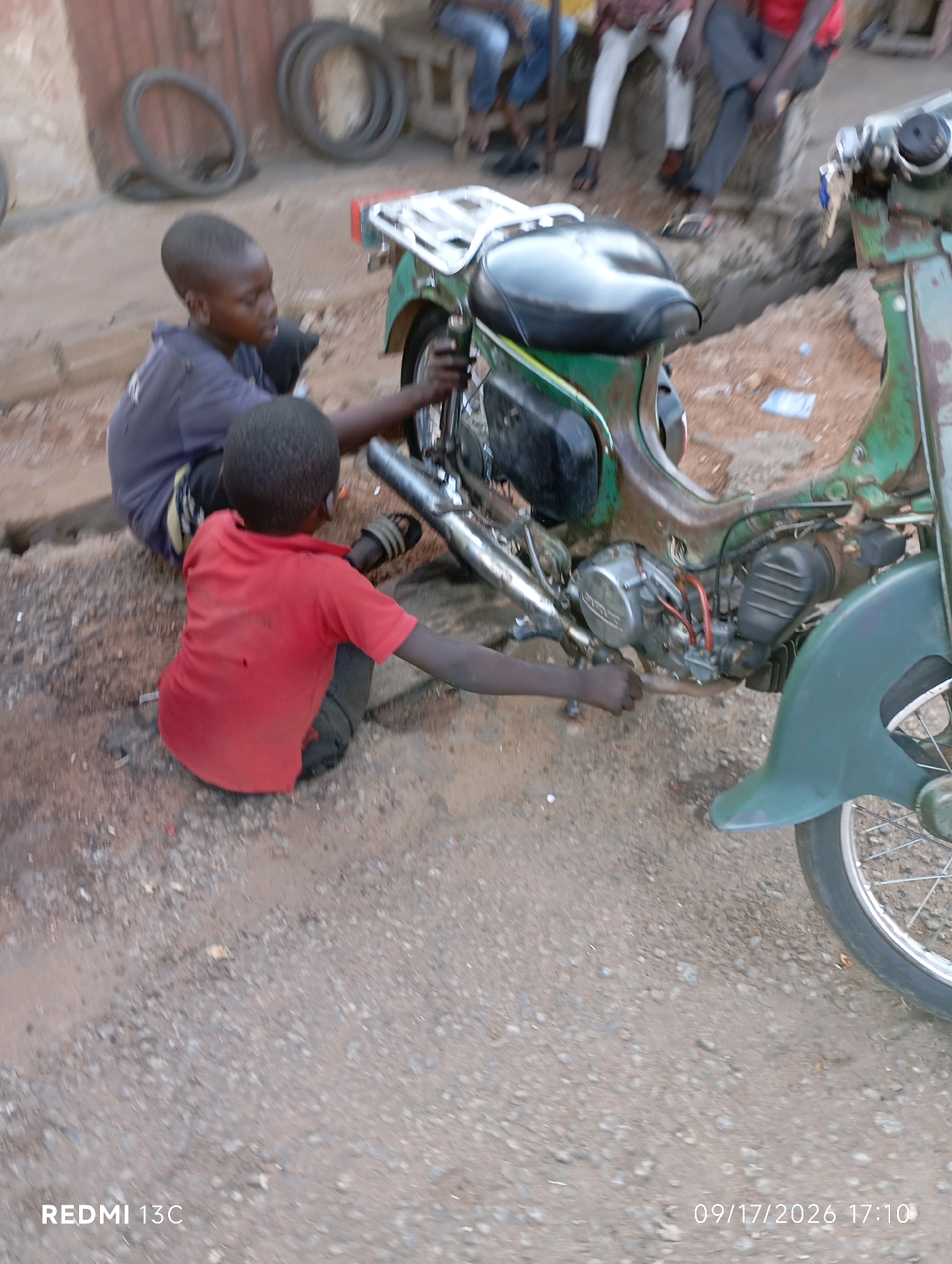 In Shendam, Nigeria, two young boys are engaged in working on a green motorcycle. One boy, wearing a dark blue shirt, is positioned behind the motorcycle, his hands near the back wheel, possibly inspecting or repairing it. The other boy, dressed in a red t-shirt, is seated on the ground in front of the motorcycle and is also interacting with its components, reaching towards the engine area with his right hand. The motorcycle appears to be an older model, with visible parts of the engine and exhaust system. The environment is a dusty, unpaved area, typical of a roadside or open lot in a town, with discarded tires visible in the background. The lighting suggests it is daytime.